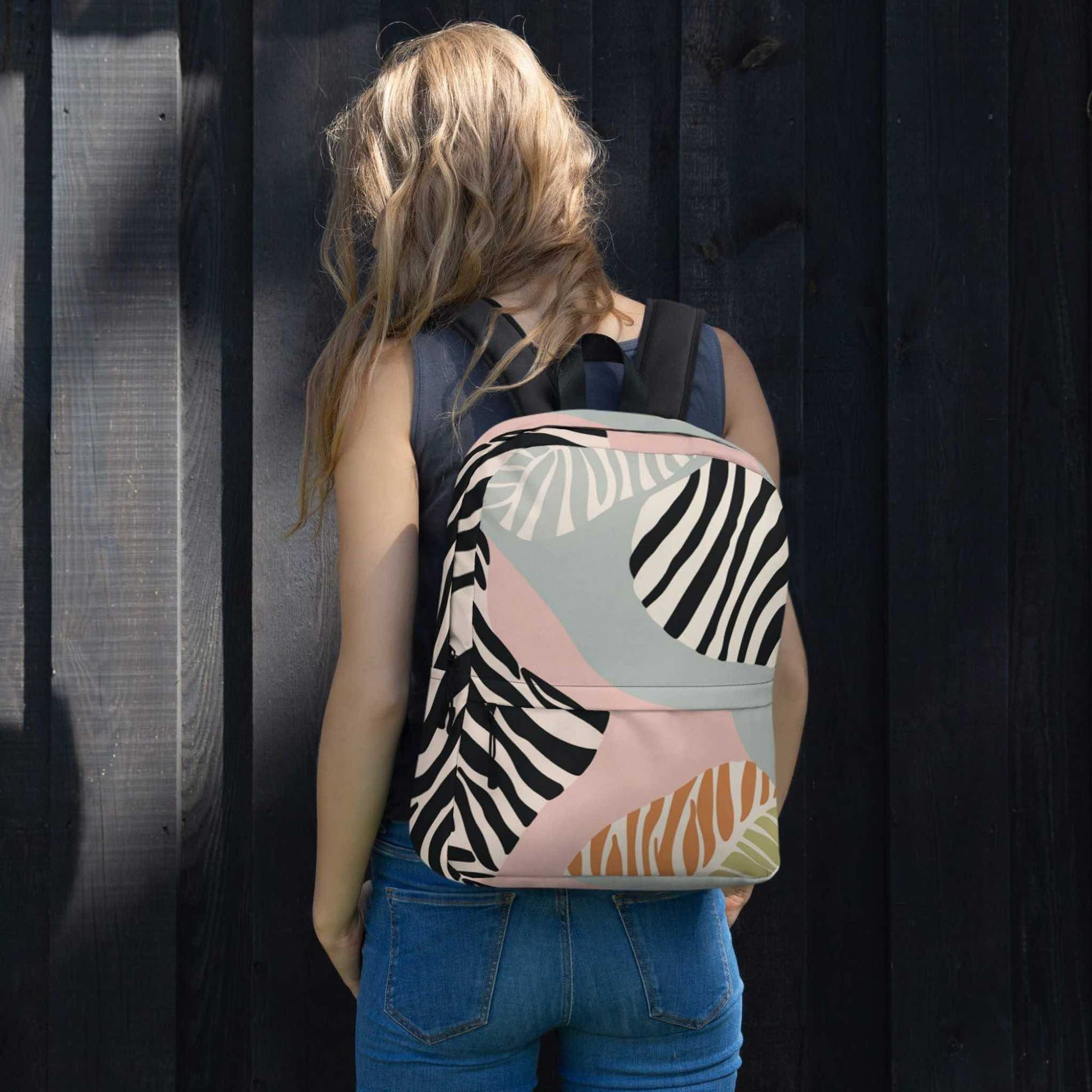 Rear view of a woman wearing a backpack which features a zebra inspired design with black, soft green, soft pink and tan colours. The woman is facing a grey feature wall.