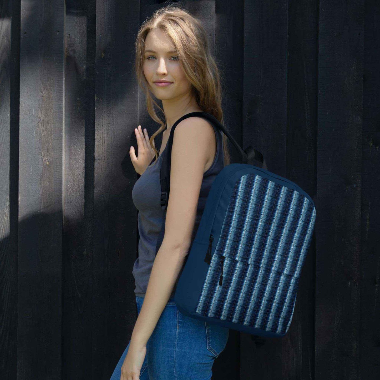 Caucasian woman with a denim stripe backpack slung over her left shoulder, looking left to the camera. The woman stands in front of dark grey feature wall.