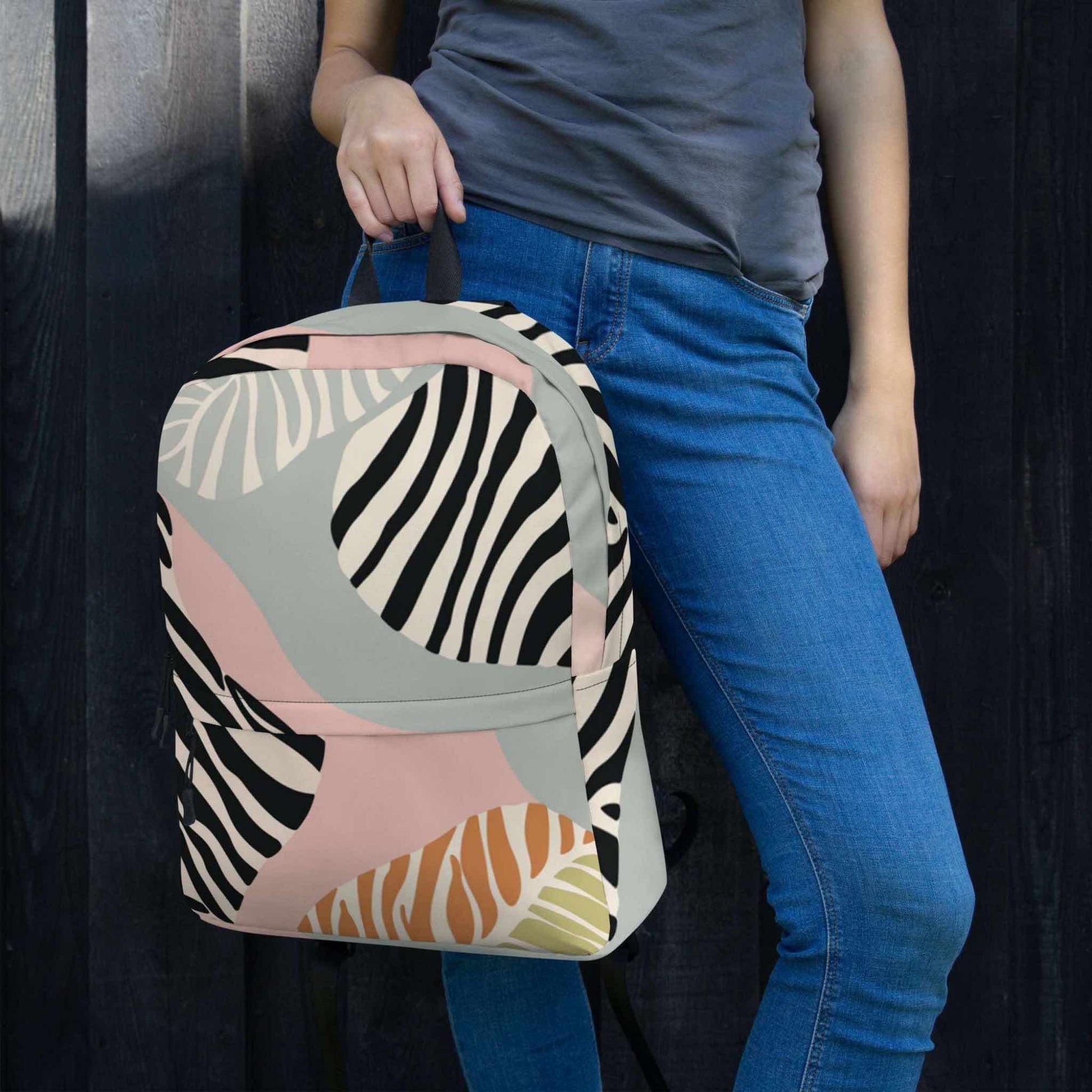 A backpack held by its top loop by a woman wearing jeans. The backpack features a zebra inspired design with black, soft pink, soft green and tan colours. The woman stands next to a dark feature wall.