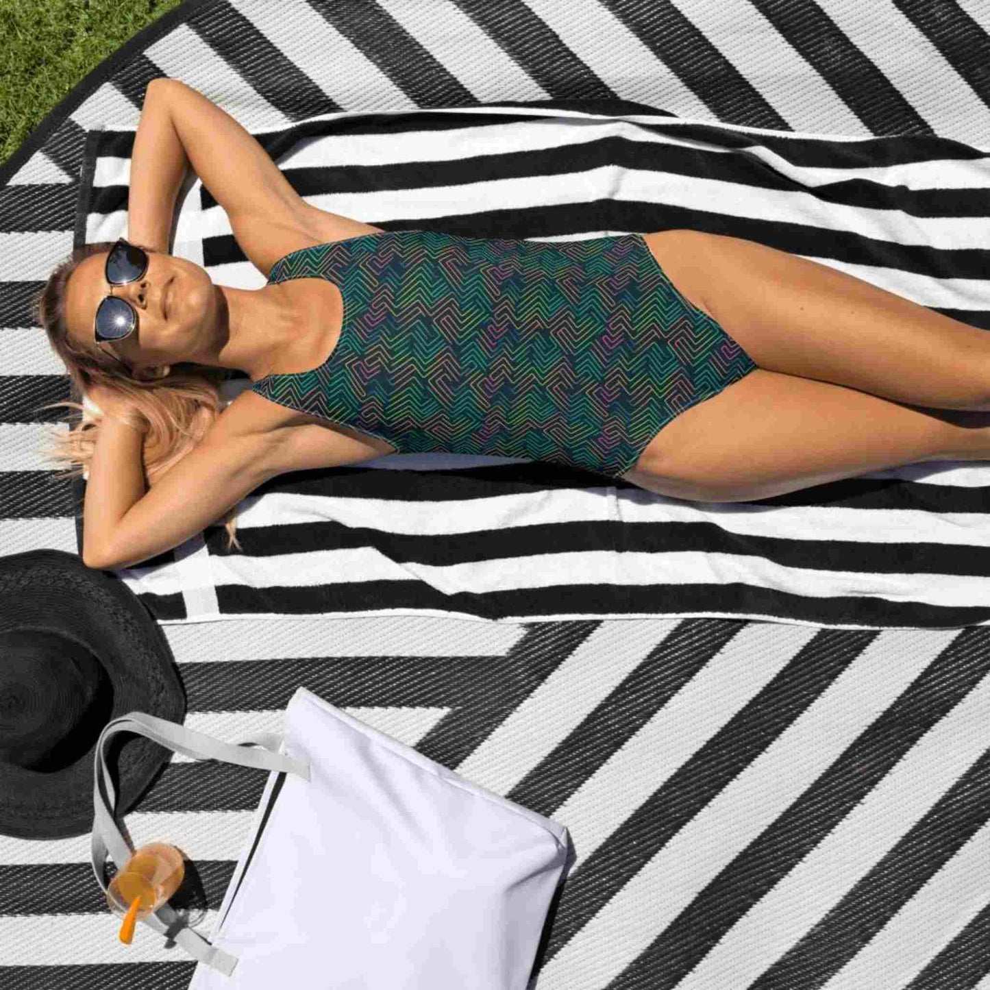 Caucasian lady with sunglasses ,in a blue one piece swimsuit with dark green neon pattern  lying face up on a beach mat in the sun..