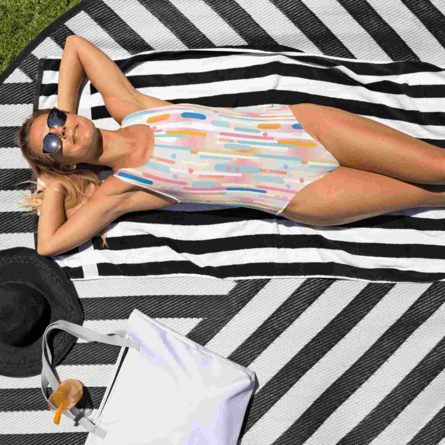 Caucasian lady with sunglasses ,in a light candy coloured one-piece swimsuit, lying face up on a beach mat in the sun.