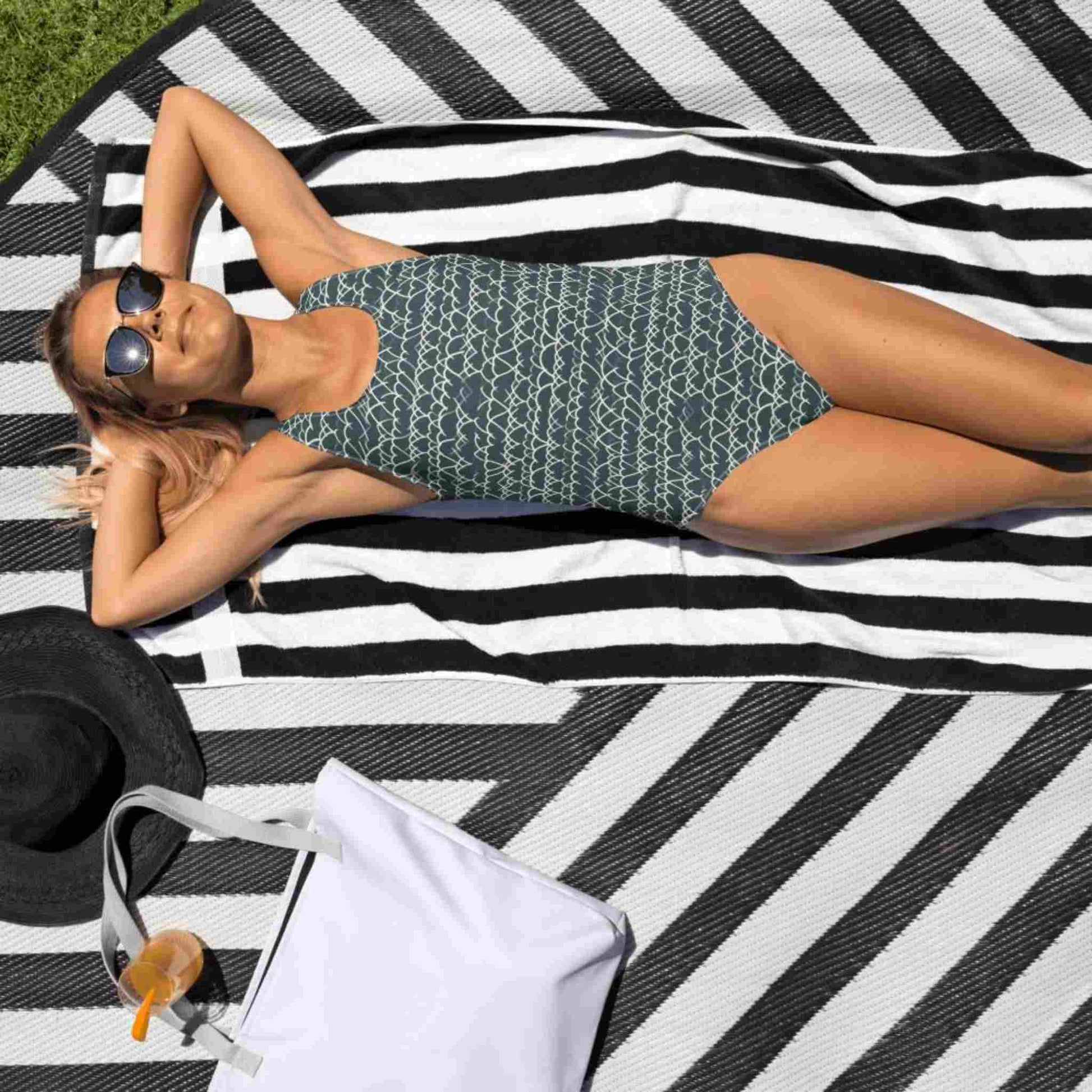 Caucasian lady with sunglasses ,in a one-piece swimsuit which features a white geometric white design that dances across a blue background, lying face up on a beach mat in the sun.