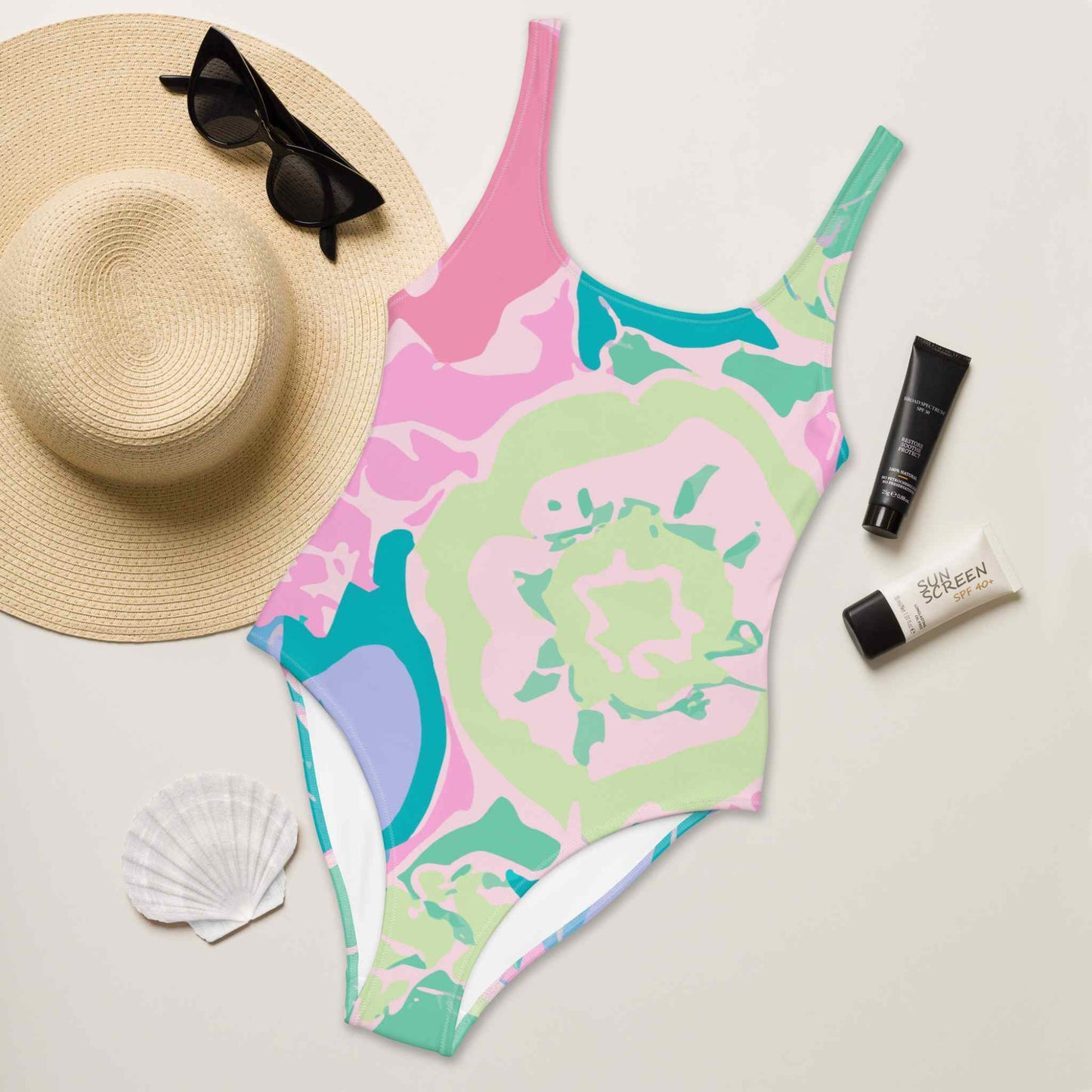 A flat lay of a one piece swimsuit, with the front of the swimsuit, in colourful pastel shades and design inspired by roses, facing up. A hat, sun screen lotions and seashell lie next to it. White background.