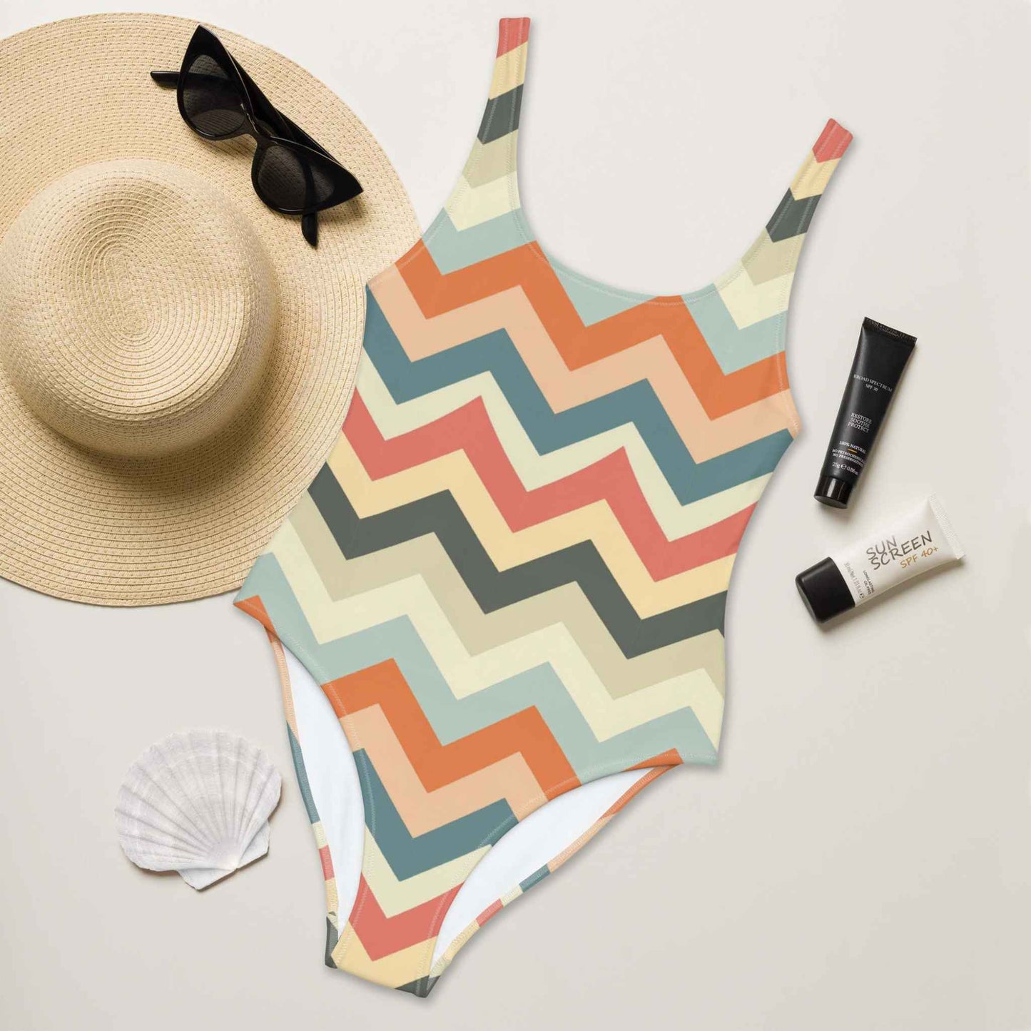 A flat lay of a one piece swimsuit, with front side facing up, designed in a chic zig zag pattern in pastel shades. A hat, sunglasses, seashell and tubes of sun screen creams laying next to it.