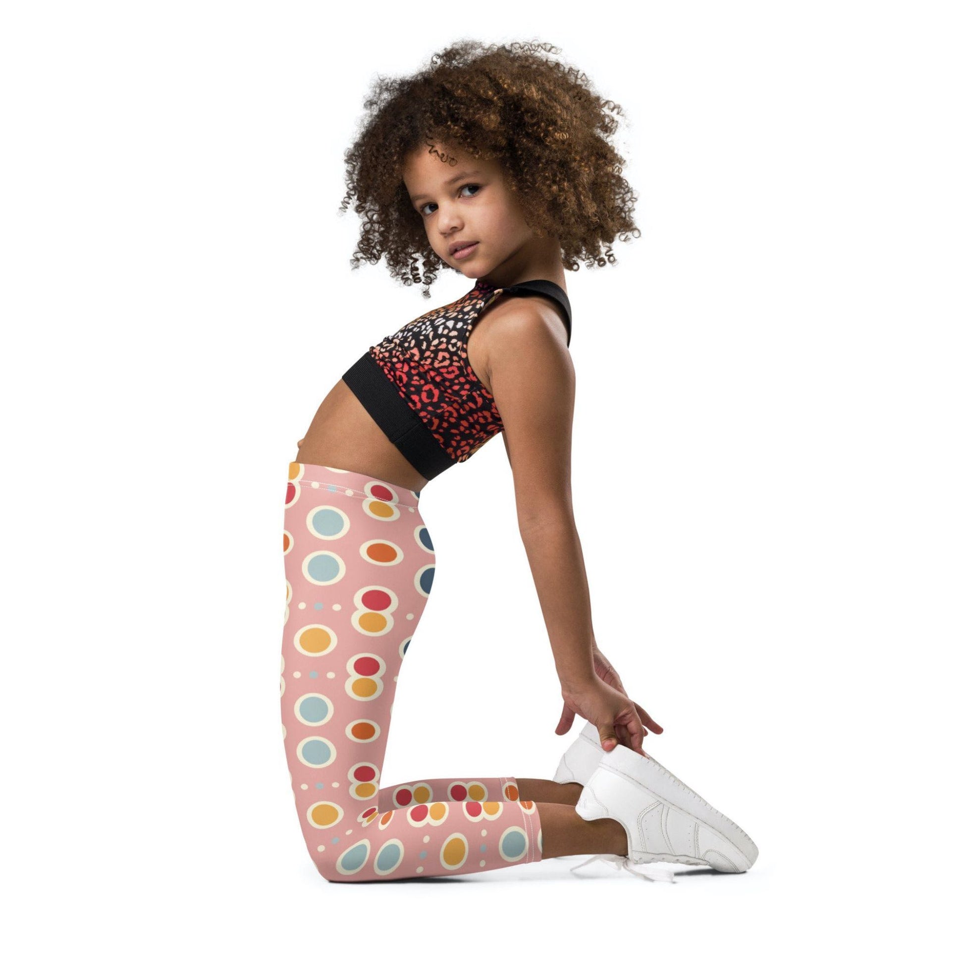A black gril wearing a crop top and leggings which features a playful pattern of pastel and white circles on a pink background. The girl is kneeling and stretched backwards as she looks left into the camera.