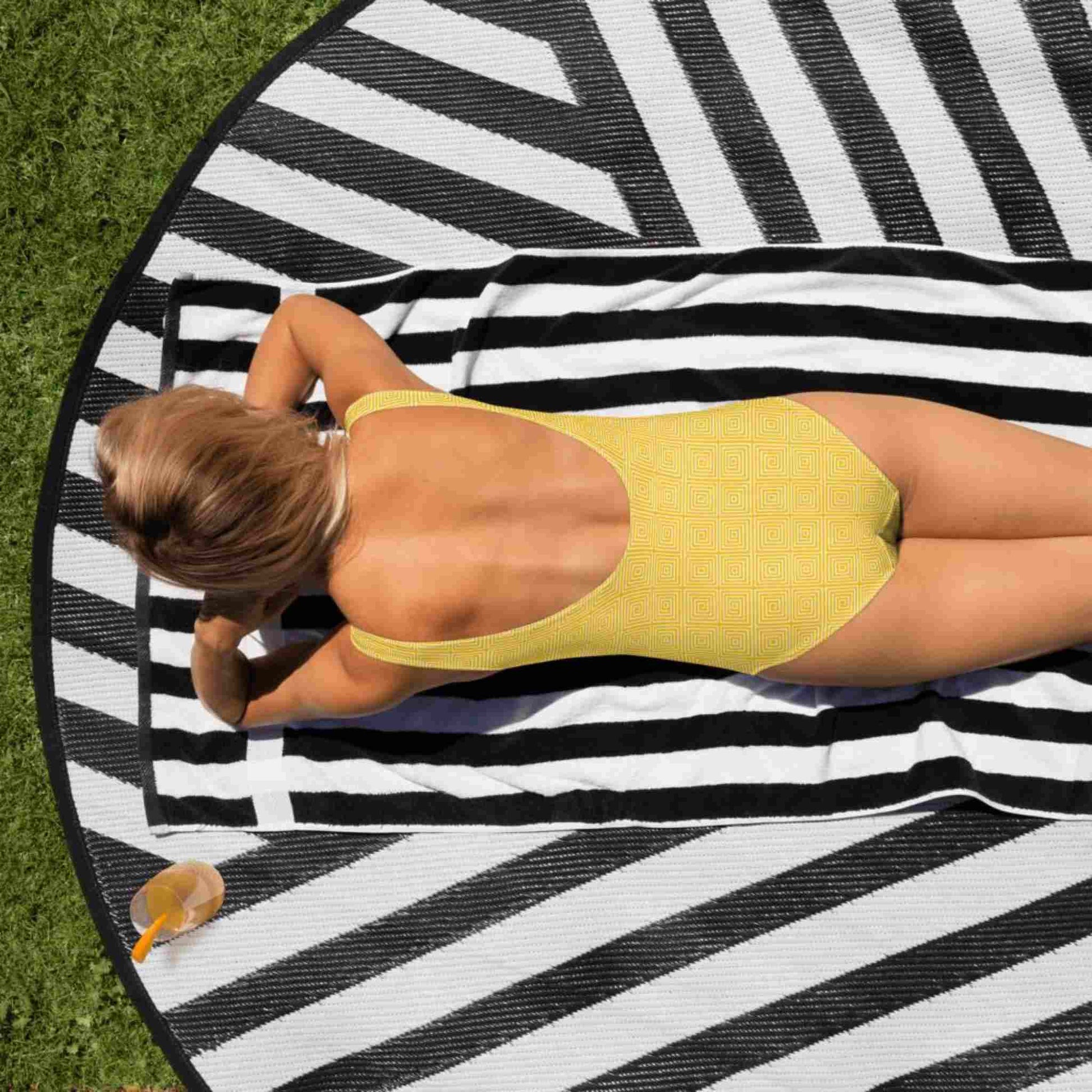 A caucasian lady in a one-piece swimsuit, which features a yellow op art illusion pattern, lying face down on a beach mat in the sun.