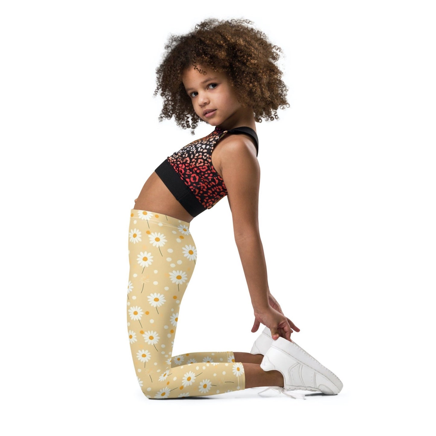 2T yellow leggings named DAISYDOODLE for toddlers, and bigger sizes for kids. This design features playful daisies &amp; polka dots on a bright egg yellow background, worn by a young girl who kneels leaning bakwards, her side of body and face facing the camera. White background.