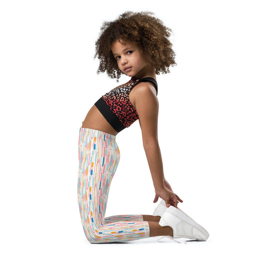 Kid and roddler leggings which features playful candy colored lines in a white background, worn by a young girl. The girl kneels and stretches backwards, in a side view to camera, her face lited left towards the camera. White background.