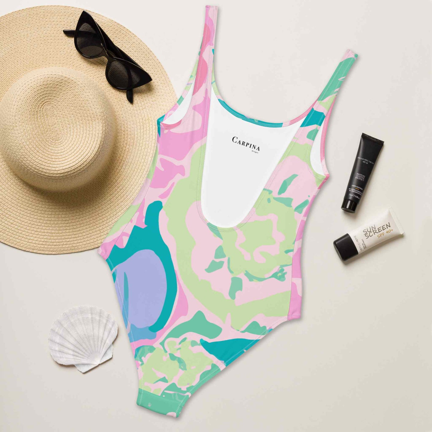 A flat lay of a one piece swimsuit, with the rear of the swimsuit, in colourful pastel shades and design inspired by roses, facing up. A hat, suncreen lotions and seashell lie next to it. White background. The Carpina Designs prnted in the interior side of the swimsuit is visible. White background.