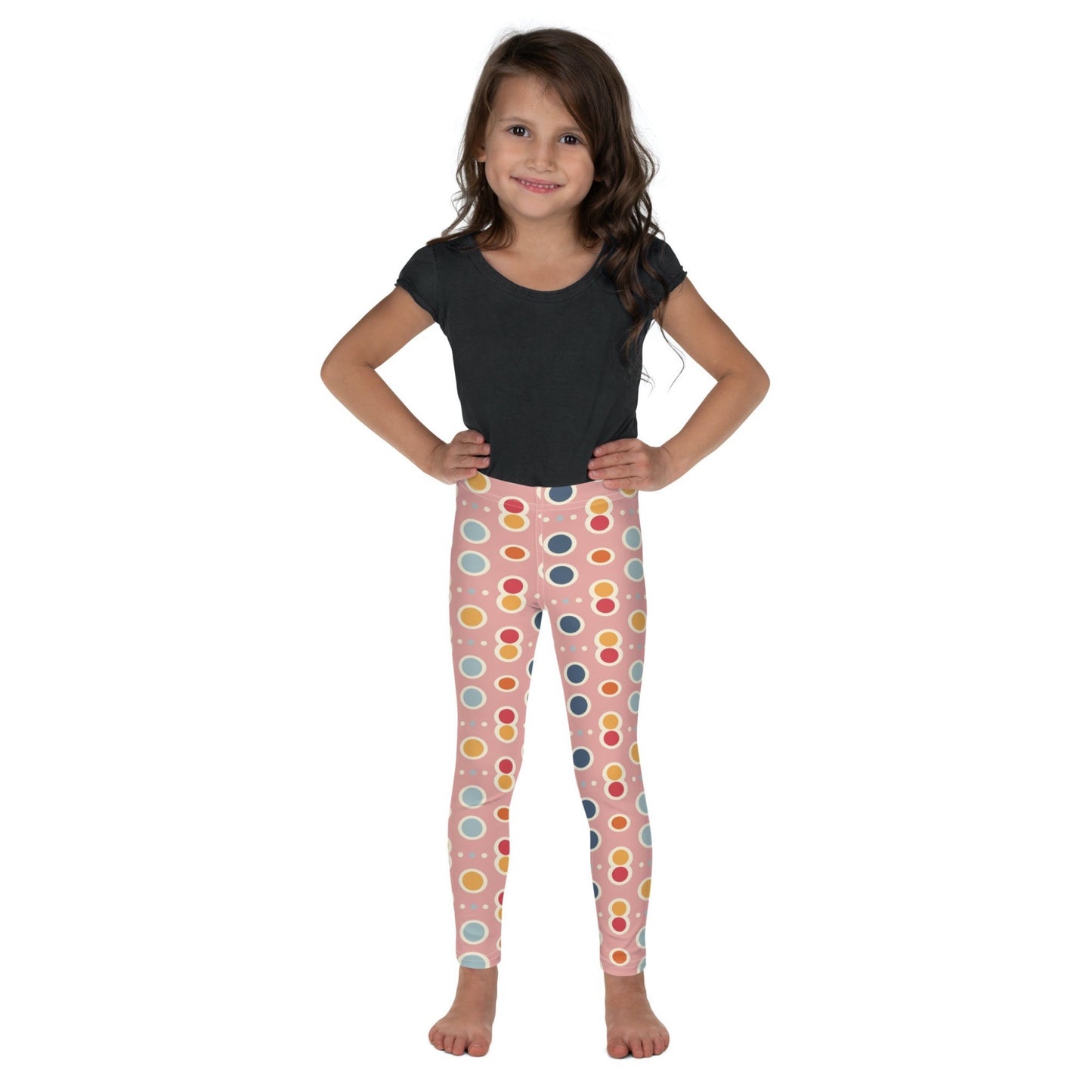 Front view of a dark skinned gril wearing a black tshirt top, and leggings which features a playful pattern of pastel and white circles on a pink background.  The girl has her arms folded on her side and is standing against a white background.