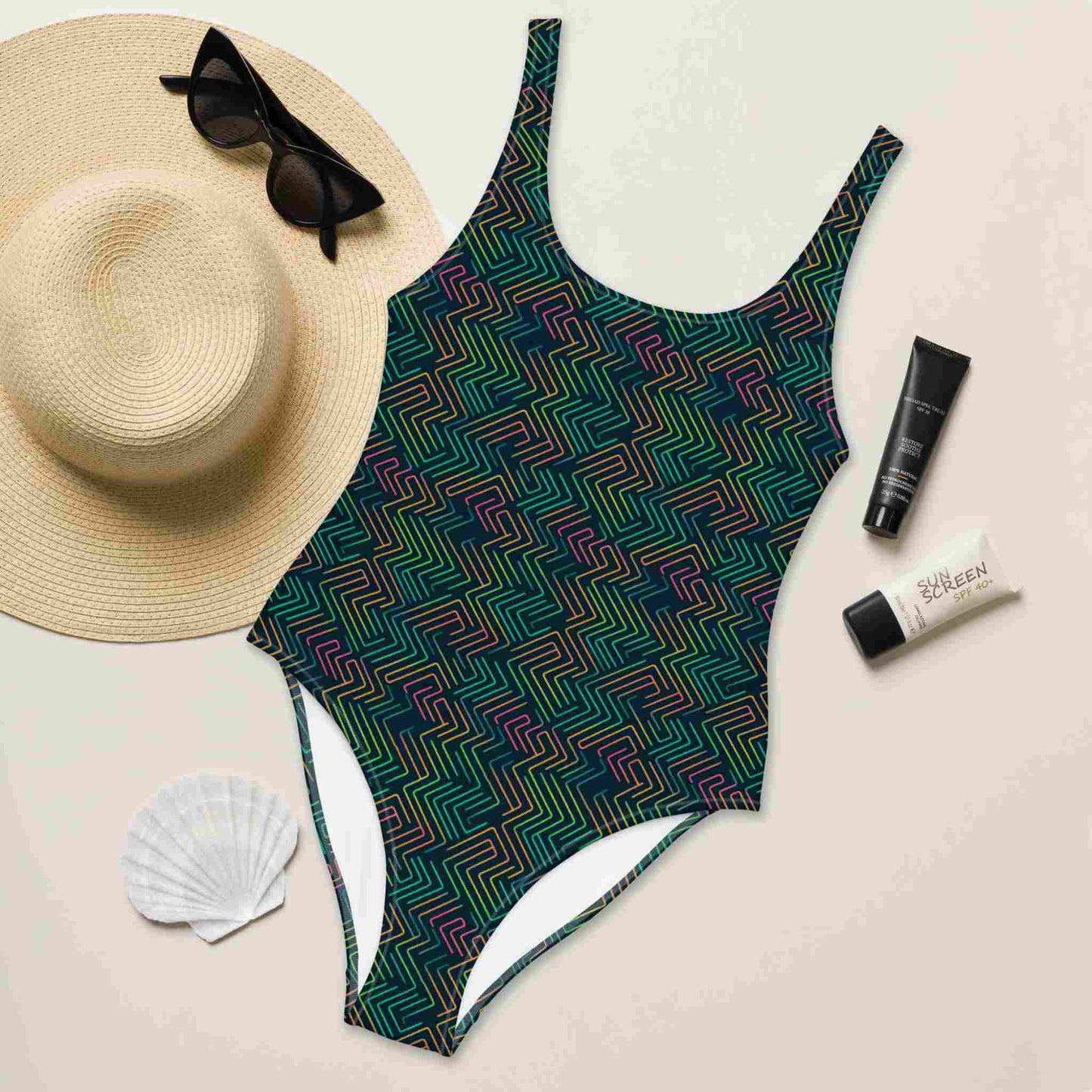 A flat lay of a blue one-piece swimsuit, in a design with dark green neon pattern, front side facing up. A hat, sunglasses, seashell and tubes of sun screen creams lying next to it.