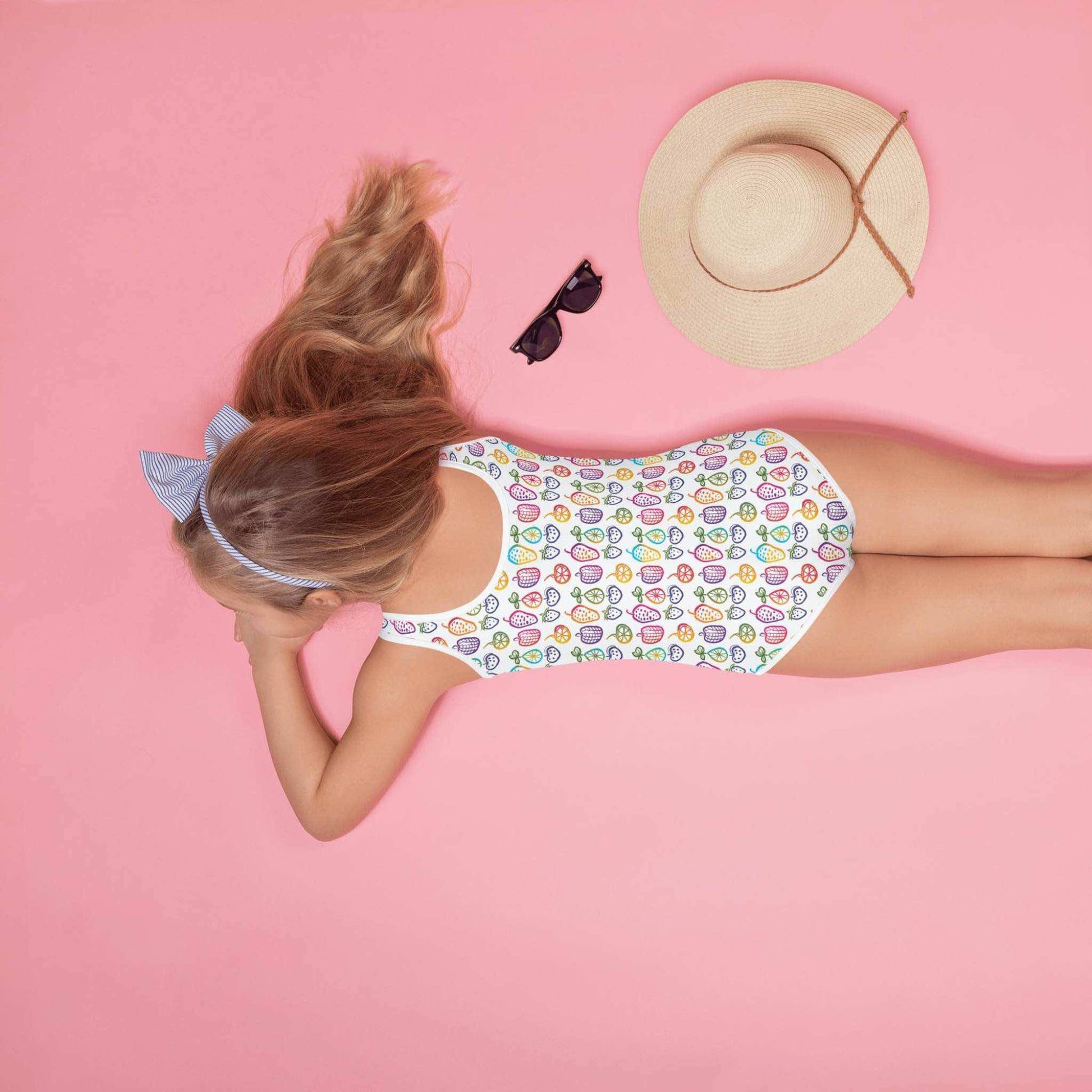 A little girl, lying face down against a pink background, wearing an adorable junior one-piece swimsuit, which features a cheerful pattern of colorful fruit doodles across a pristine white background.