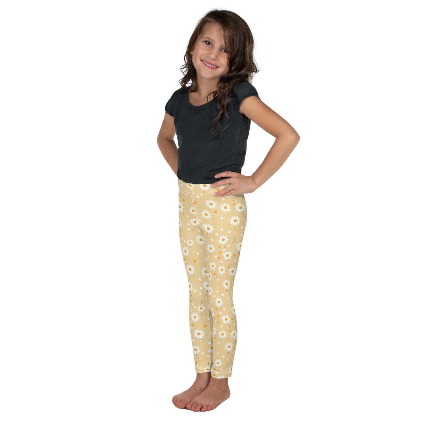 Frontal left view of a smiling dark skinned gril wearing a black tshirt top, and leggings which feature sweet daisy prints on an egg yellow background. The girl has her arms folded on her side and is standing against a white background.