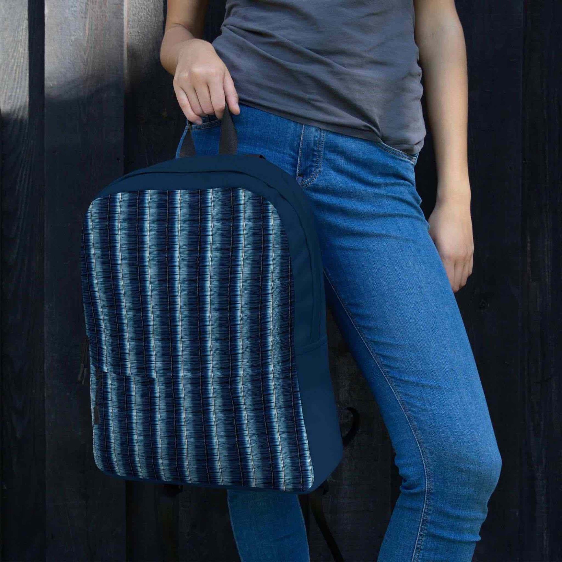 A denim stripe backpack held by its top loop by a woman wearing jeans. The woman stands next to a dark feature wall.