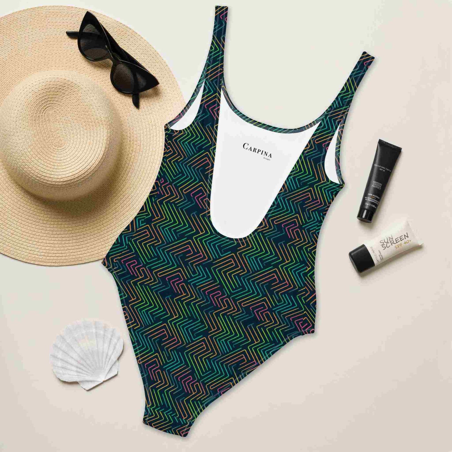 A flat lay of a blue one-piece swimsuit, in a design with dark neon green pattern, rear side of swimsuit facing up and revealing the Carpina Design inside label. A hat, sunglasses, seashell and tubes of sun screen creams lying next to it.