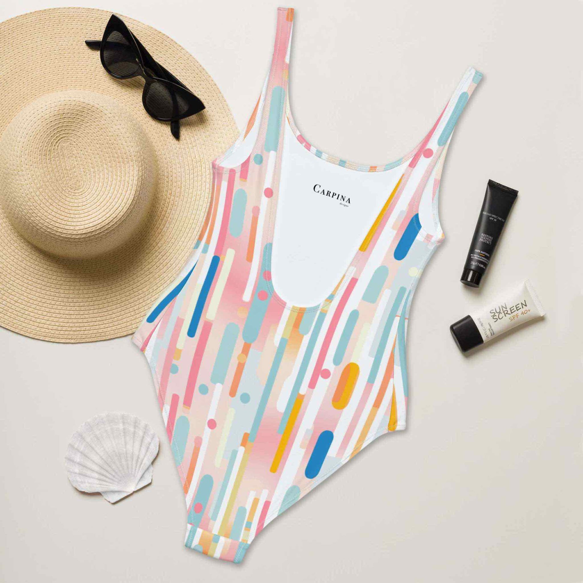 A flat lay of a light candy coloured one-piece swimsuit, back side of swimsuit facing up and revealing the Carpina Design inside label. A hat, sunglasses, seashell and tubes of sun screen creams lying next to it.