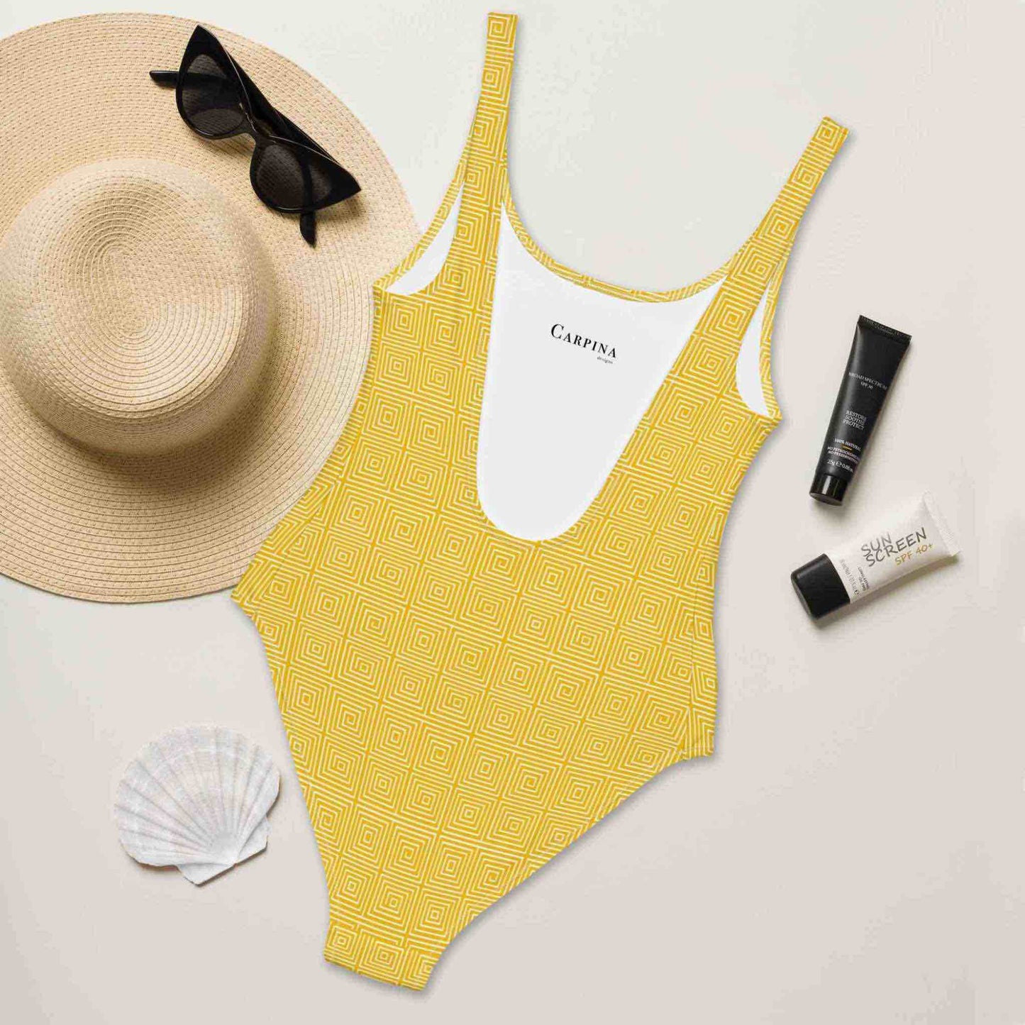 A flat lay of a one-piece swimsuit, which features a yellow op art illusion pattern, rear side of swimsuit facing up and revealing the Carpina Design inside label. A hat, sunglasses, seashell and tubes of sun screen lie next to it.