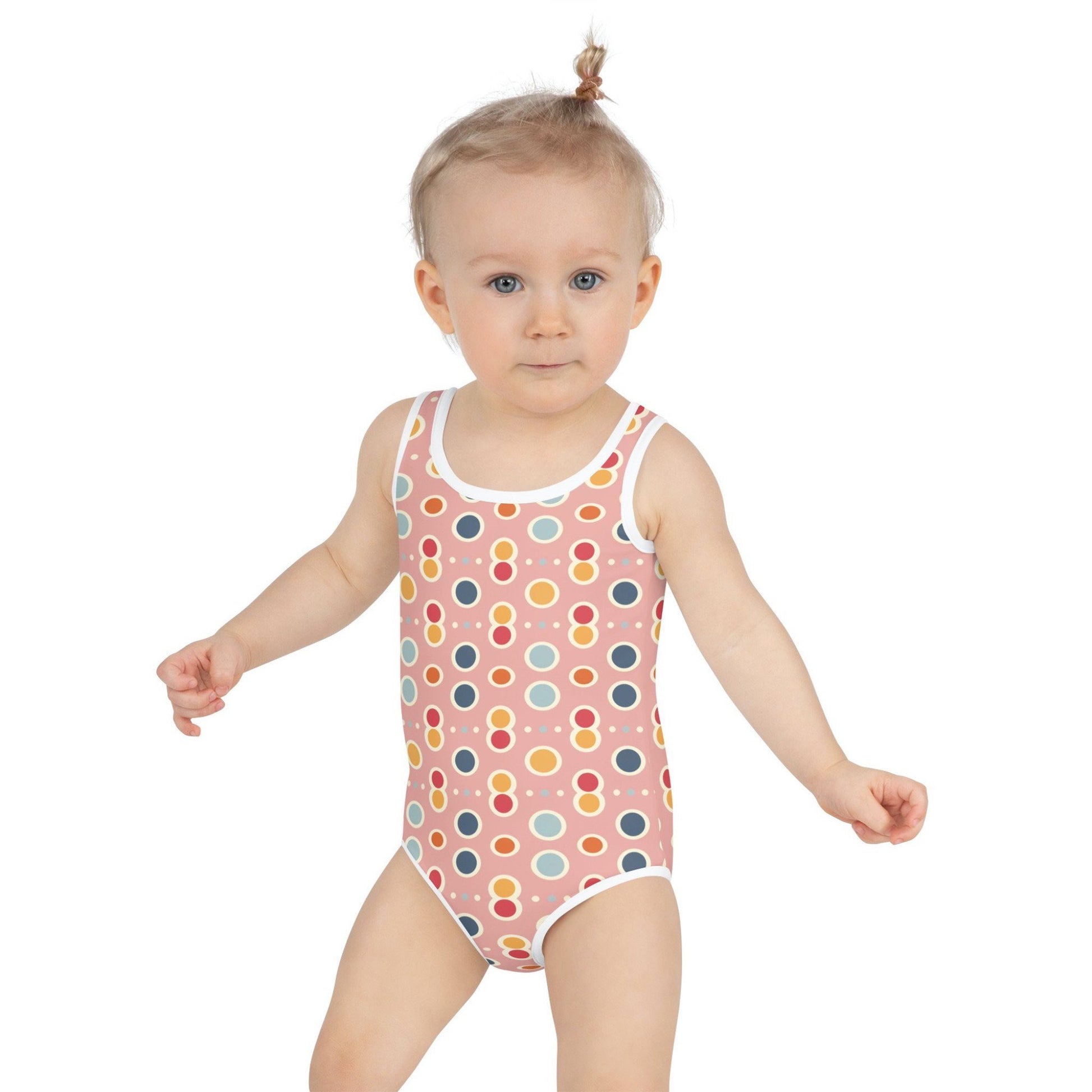 Frontal view of an infant girl wearing an adorable junior swimsuit which features a playful pattern of pastel pops, white outlines on a pink background, against a white background.