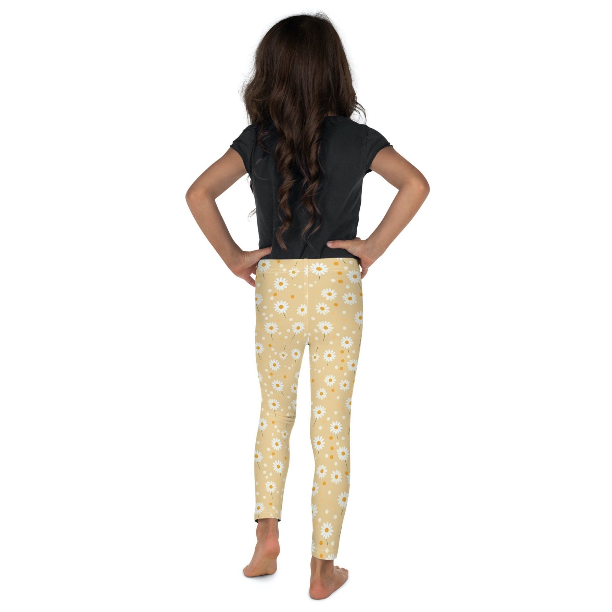 Back view of a dark skinned gril wearing a black tshirt top,  and leggings which feature sweet daisy prints on an egg yellow background. The girl has her arms folded on her side and is standing facing a white background.