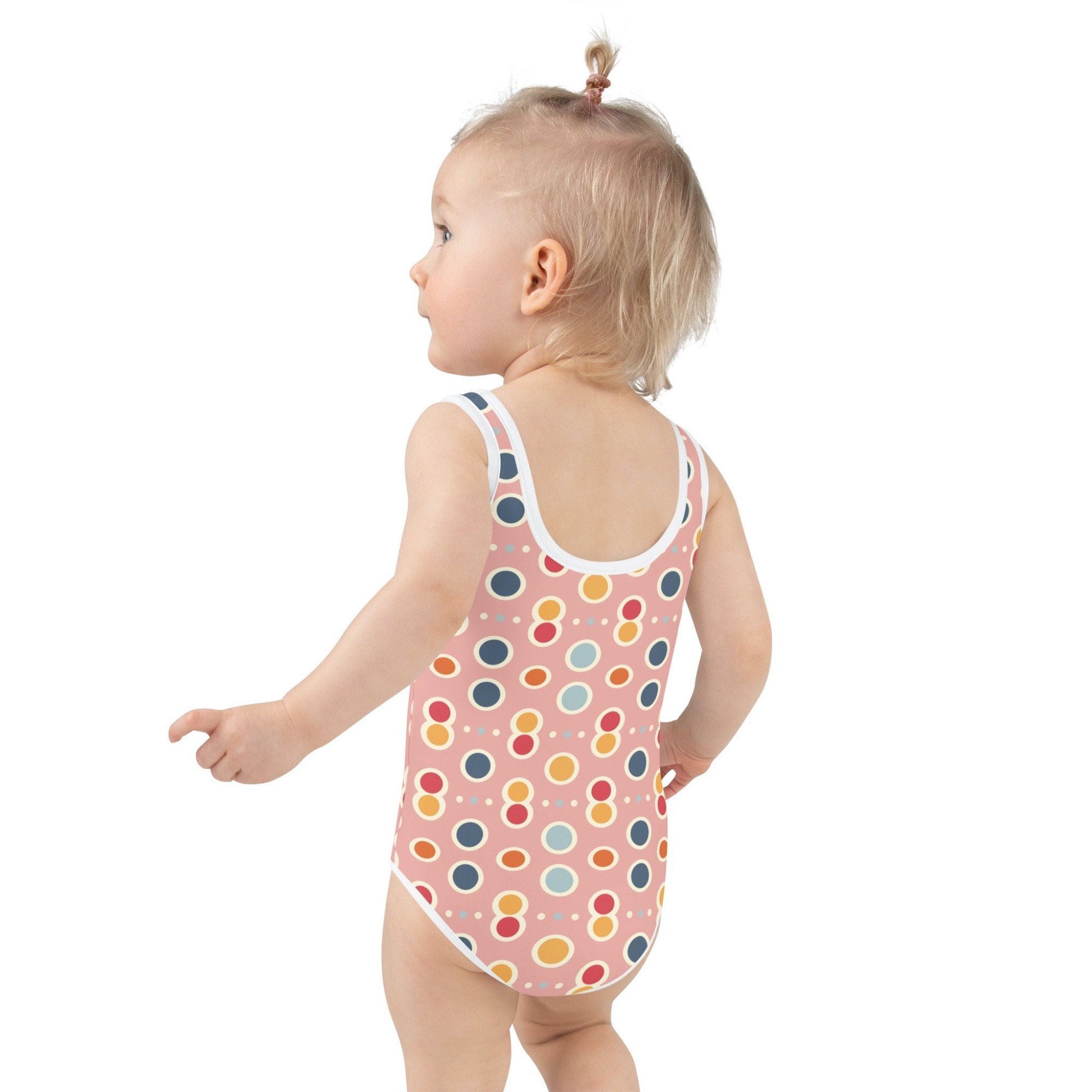 Left side rear profile of an infant girl wearing an adorable junior swimsuit which features a playful pattern of pastel pops, white outlines on a pink background, against a white background.