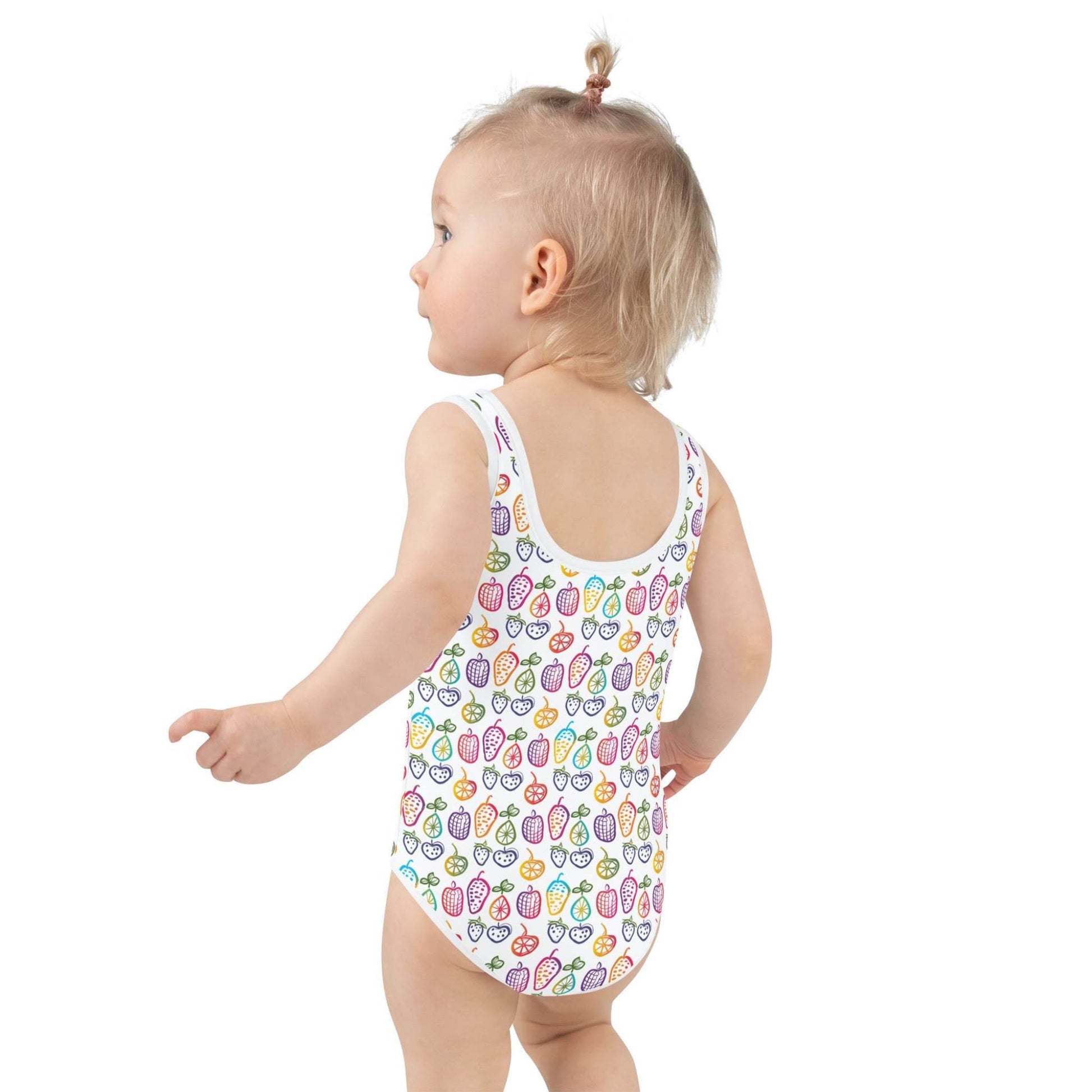 Left side rear profile of an infant girl wearing an adorable junior one-piece swimsuit which features a cheerful pattern of colorful fruit doodles against a pristine white background.