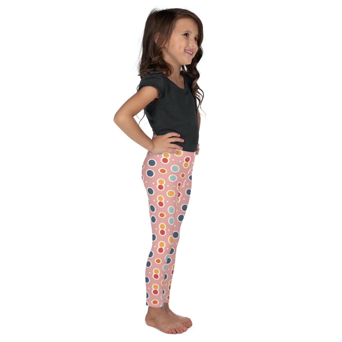 Right side view of a smiling dark skinned gril wearing a black tshirt top, and leggings which features a playful pattern of pastel and white circles on a pink background. The girl has her arms folded on her side and is standing against a white background.