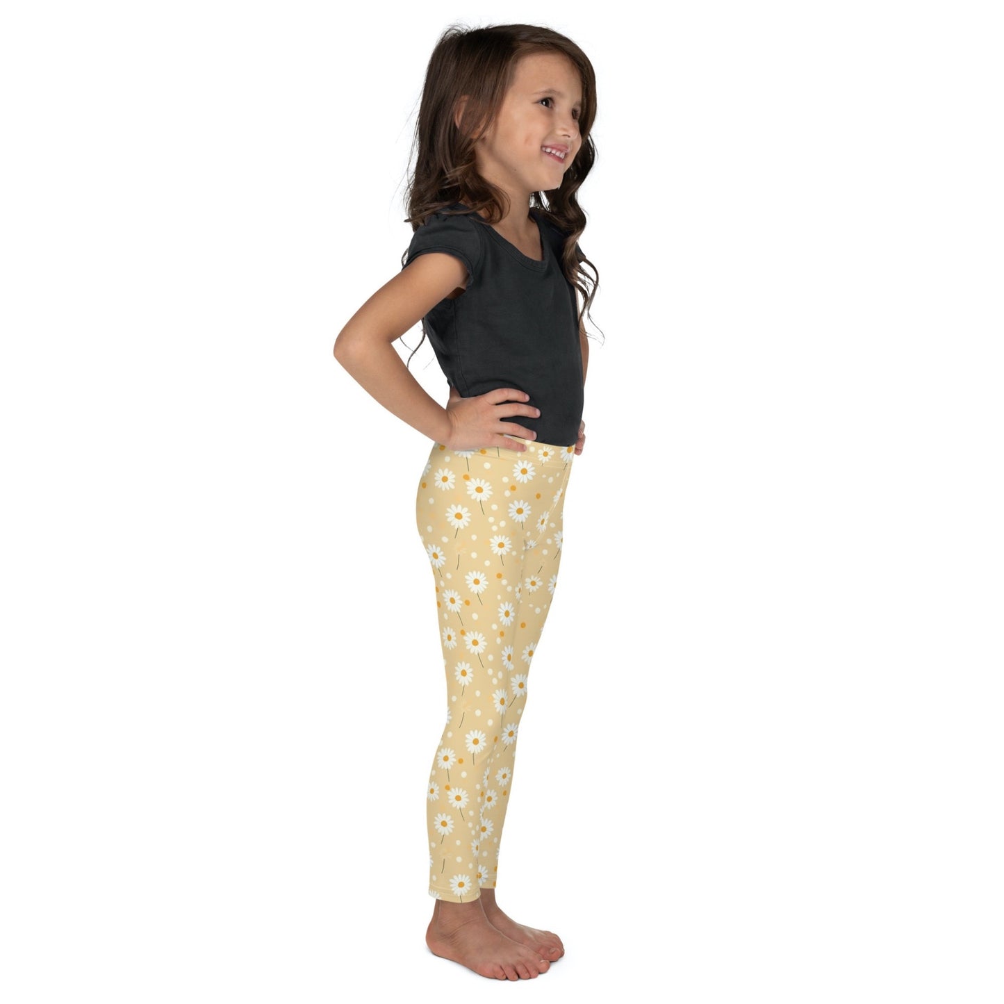 Right side view of a smiling dark skinned gril wearing a black tshirt top, and leggings which feature sweet daisy prints on an egg yellow background. The girl has her arms folded on her side and is standing against a white background.