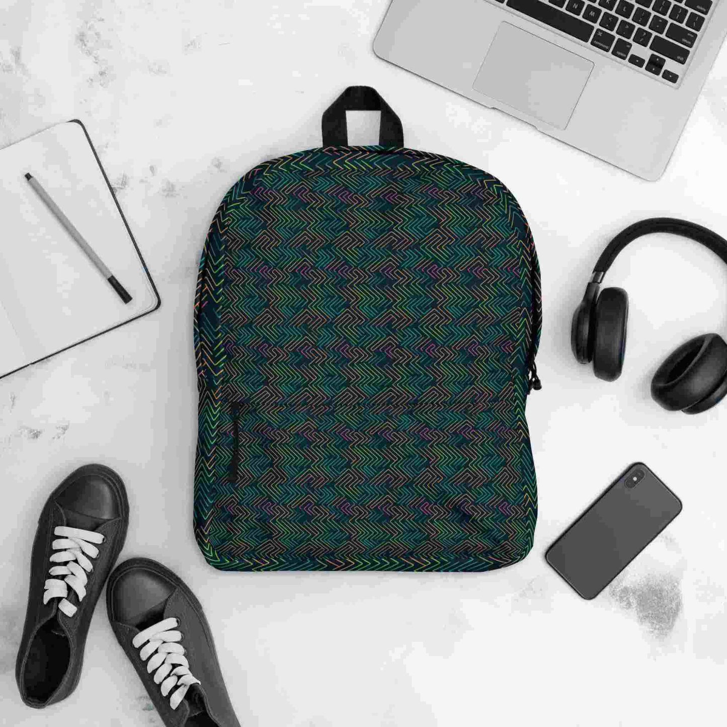 Flatlay of backpack with a dark neon pattern akin to electrical circuits, facing up, with a notepad, shows, notebook computer, earphones and mobile phone lying next to it, against a white background.