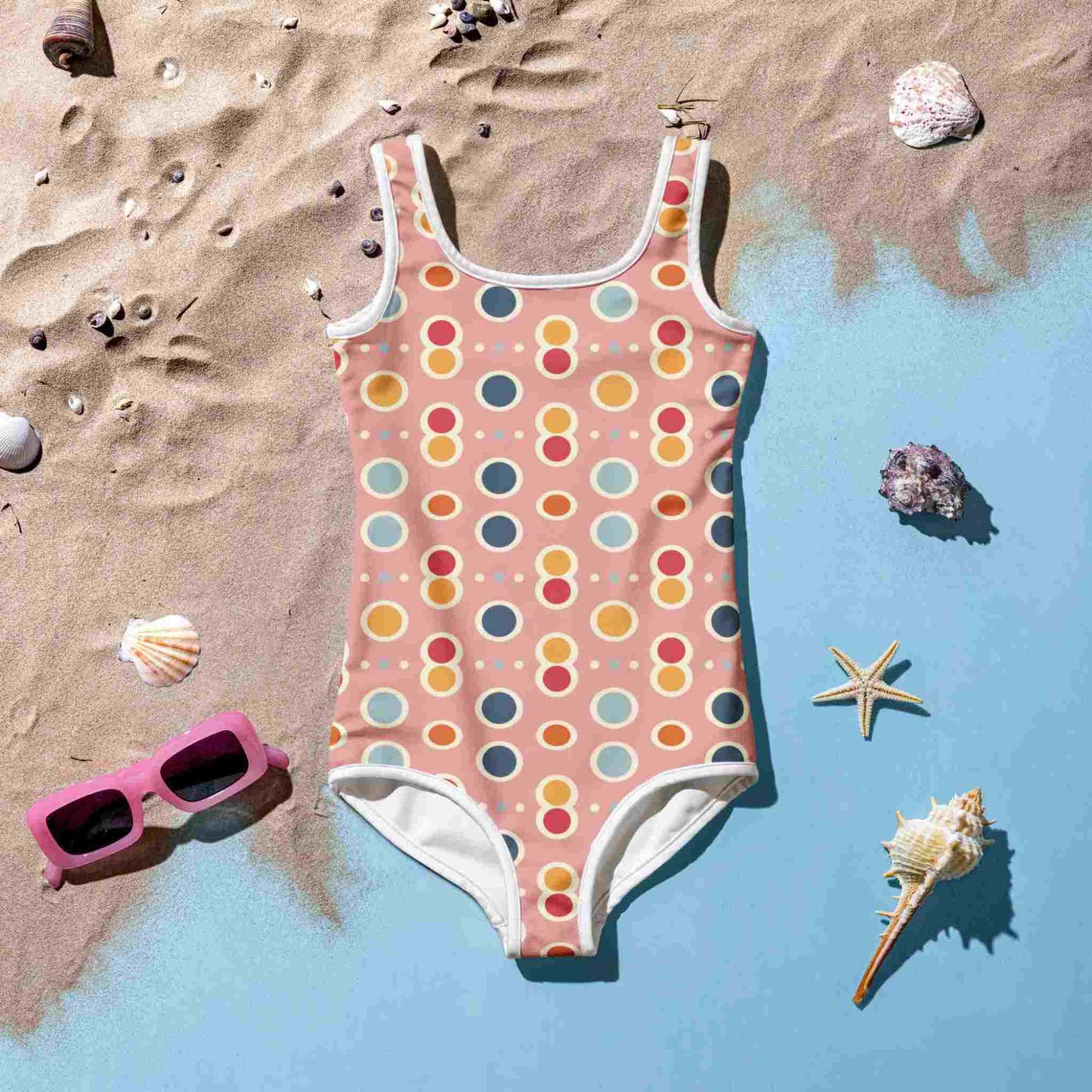 Flatlay of an adorable junior swimsuit which features a playful pattern of pastel pops, white outlines on a pink background, lying front side up on a blue floor with sea sand. A set of pink sunglasses, pebbles, starfish and sea shells lie around the swimsuit..