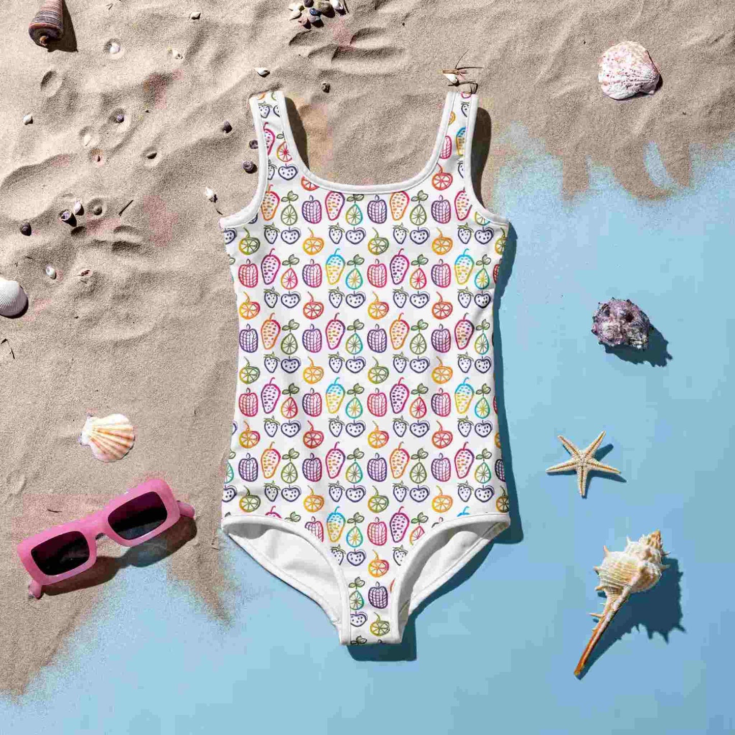 Flatlay of an adorable junior one-piece swimsuit which features a cheerful pattern of colorful fruit doodles across a white background., lying front side up on a blue floor with sea sand. A set of pink sunglasses, pebbles, starfish and sea shells lie around the swimsuit.