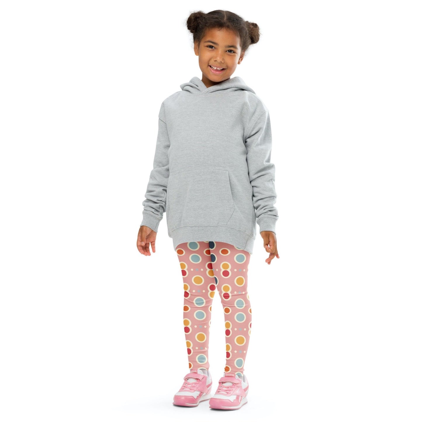 Front view of a smiling black girl whose hair is bound into buns wearing a grey sweater, and leggings which features a playful pattern of pastel and white circles on a pink background. The girl is standing against a white background.