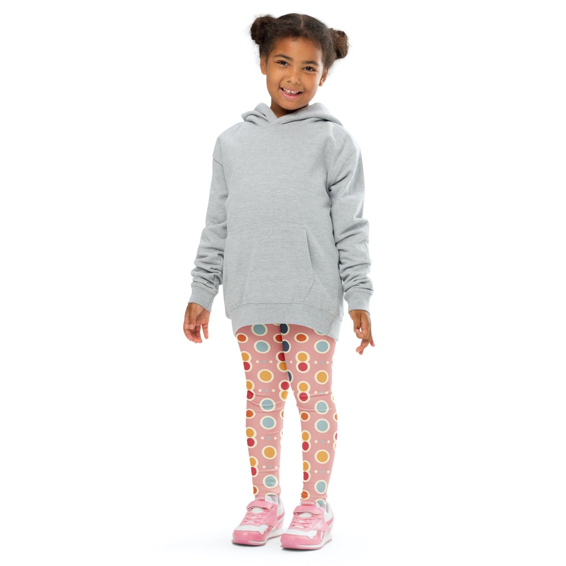 Front view of a smiling black girl whose hair is bound into buns wearing a grey sweater, and leggings which features a playful pattern of pastel and white circles on a pink background. The girl is standing against a white background.