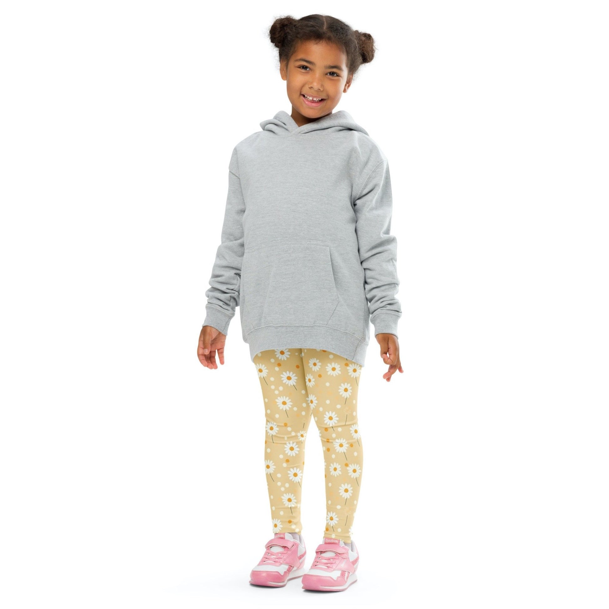 Front view of a smiling black girl whose hair is bound into buns wearing a grey sweater, and leggings which feature sweet daisy prints on an egg yellow background. The girl is standing against a white background.