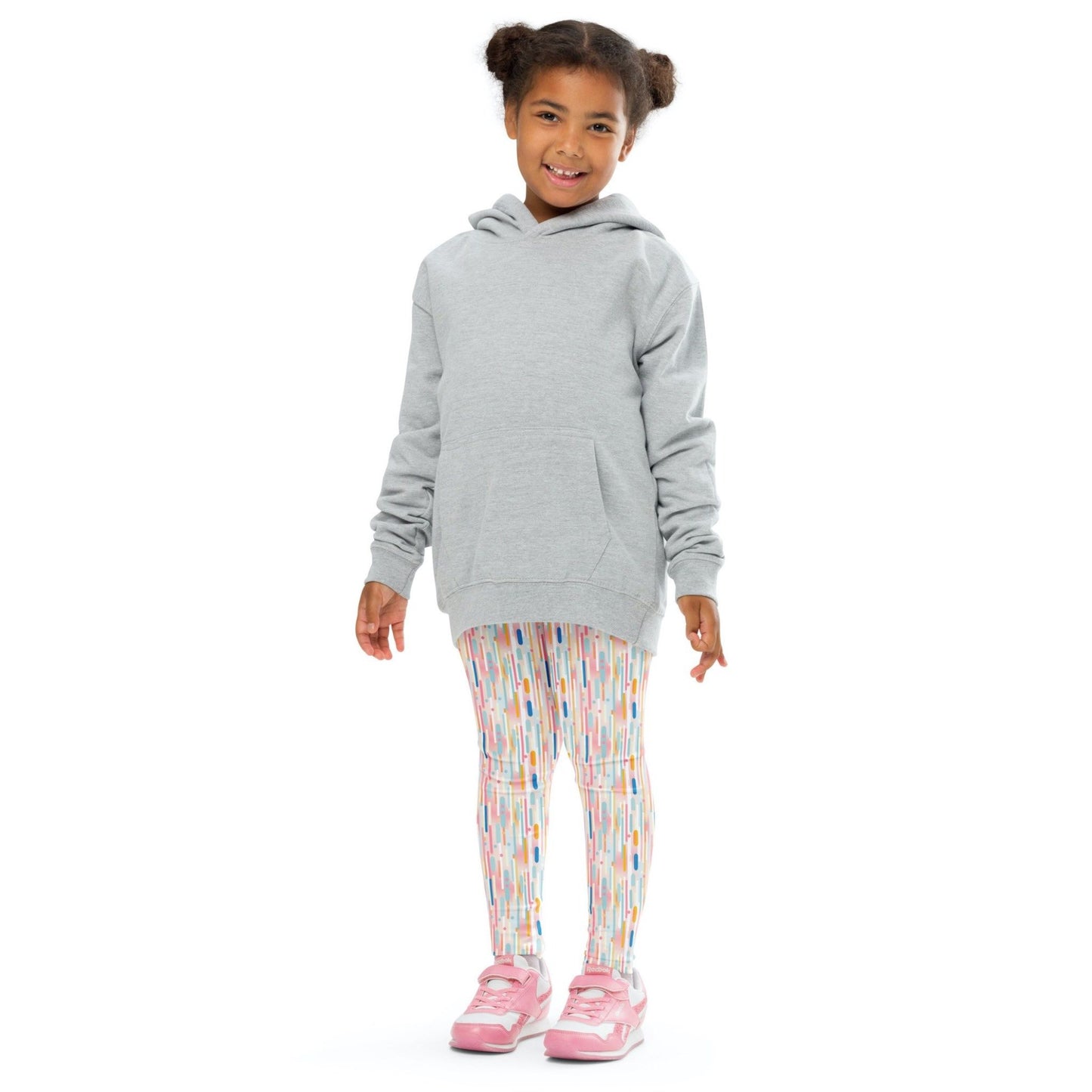 Front view of a smiling black girl whose hair is bound into buns wearing a grey sweater, and leggings which features playful candy colored lines in a white background. The girl is standing against a white background.