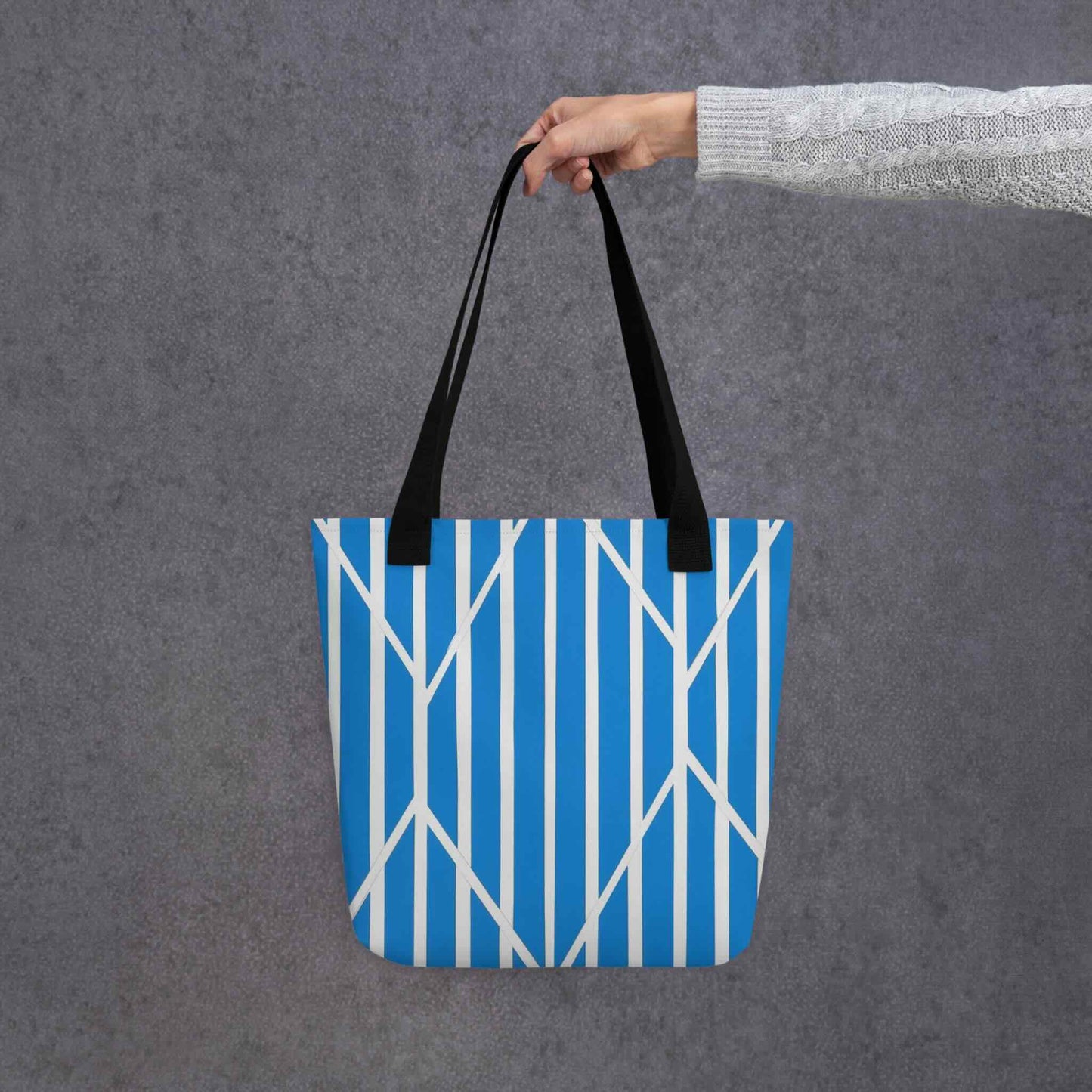 A tote bag with a striking design of blue and white striped tote which is a piece of kinetic art, held in a hand. The ordered rhythm of vertical stripes is dramatically intersected by bold, angular lines, creating a beautiful tension and dynamic focal point. Dimensions 15x15 inches. Grey background. 