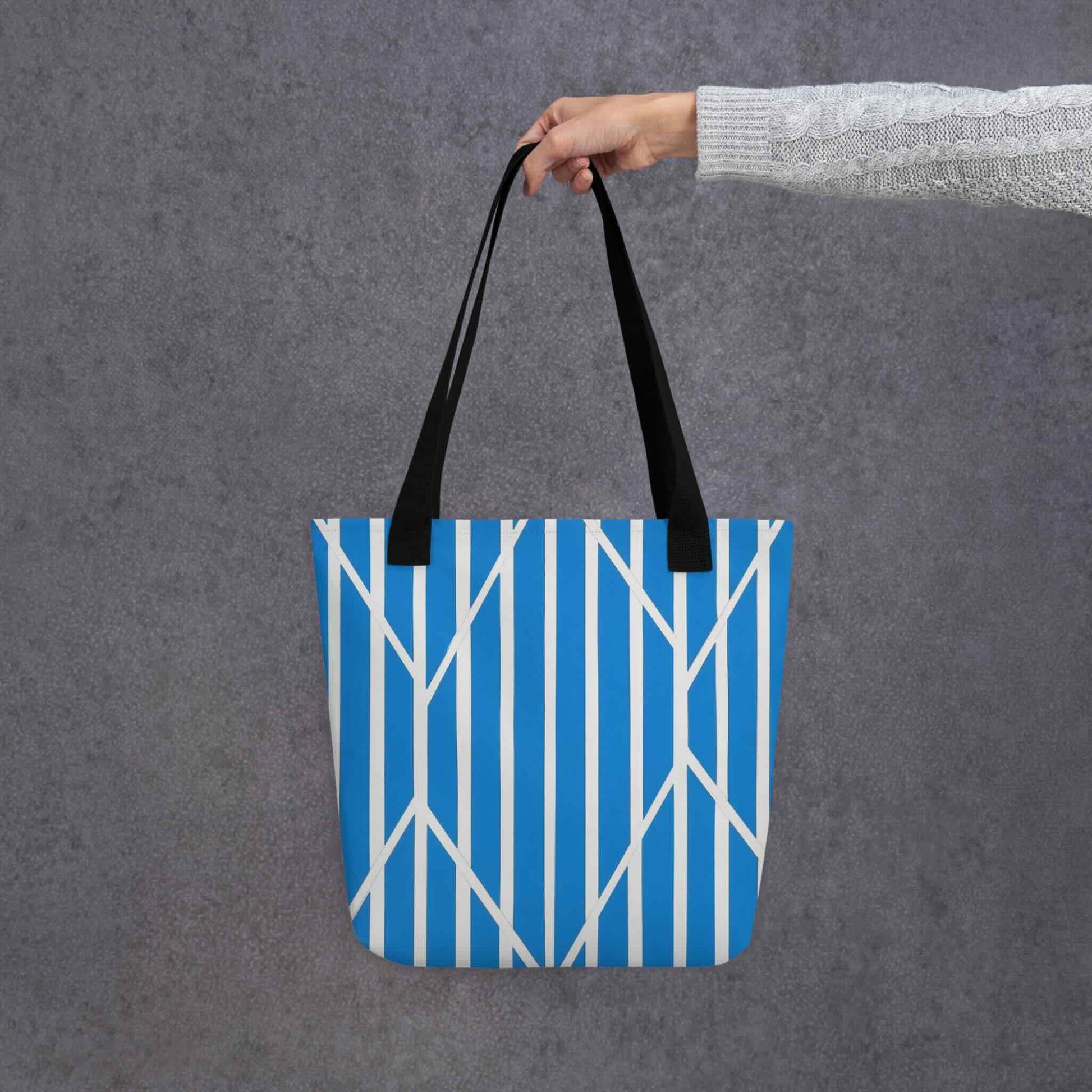 A tote bag with a striking design of blue and white striped tote which is a piece of kinetic art, held in a hand. The ordered rhythm of vertical stripes is dramatically intersected by bold, angular lines, creating a beautiful tension and dynamic focal point. Dimensions 15x15 inches. Grey background. 