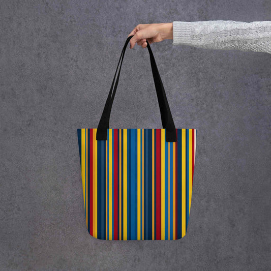 A blue striped tote bag which features a stunning geometric pattern in rich navy, glamorous gold, and deep burgundy, capturing the sophistication of high-end design, held in a hand. Black handles. Dimension 15x15 Inch polyester tote. Grey background. 