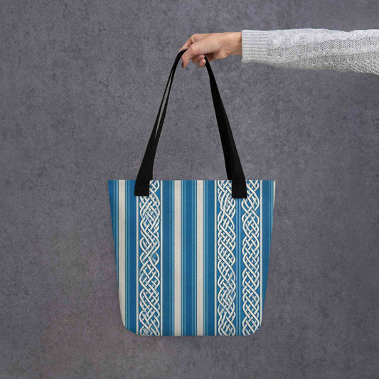 A sophisticated celtic tote bag which features intricate white knotwork columns flowing over deep marine blue and textured cream stripes, held in a hand. Grey background.