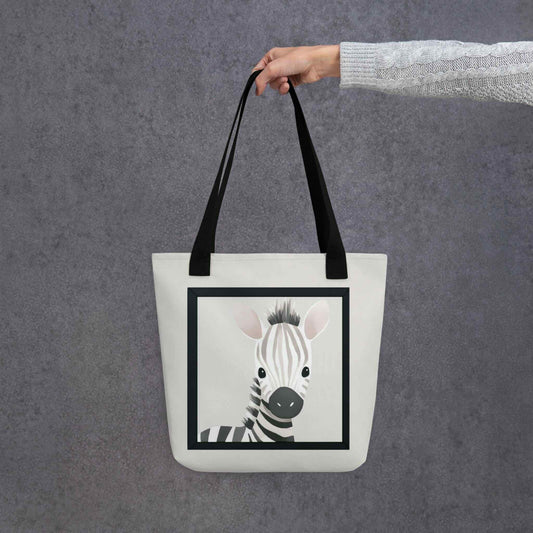 Cute totes with watercolor illustration that captures the sweet, gentle innocence of a baby zebra with soft taupe and cream stripes, topped with an irresistibly spiky mane, held in a hang. Black handles. Dimensions 15x15 inches. Grey background.
