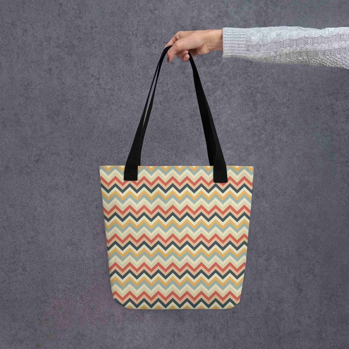 A 15x15 inch tote bag with black handles, in a multicoloured pattern of Zig Zag lines, held in a lady's hand. Grey background.