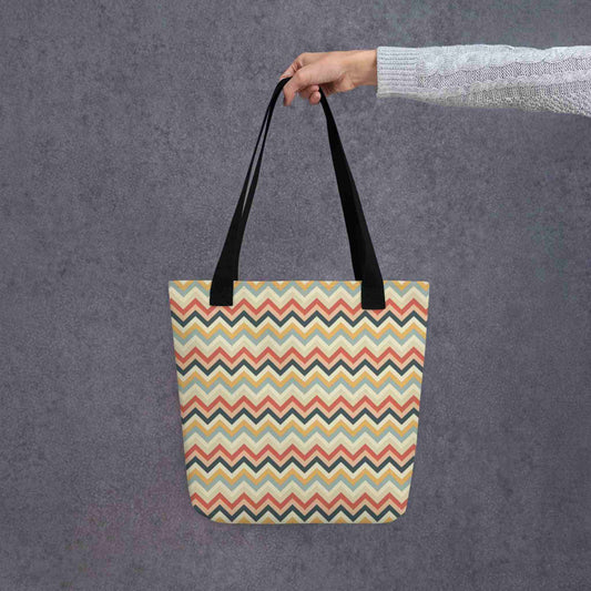 Zig zag tote bag with black handles, in a multicoloured pattern of Zig Zag lines, held in a lady's hand. Dimension 15x15 inches. Grey background.