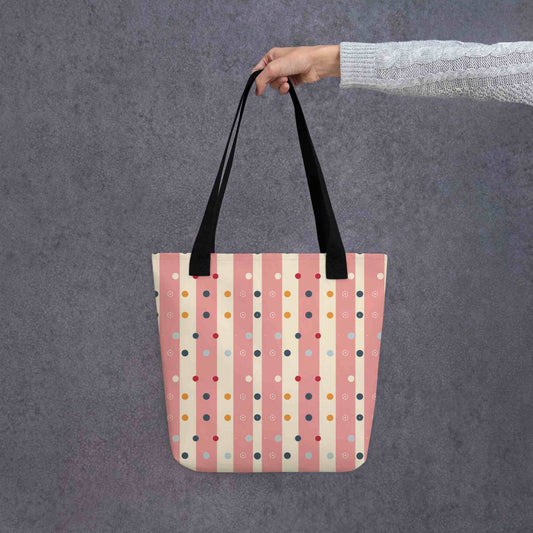 Pink striped tote bag with black handles, which features pink and beige stripes with colourful pokka dots, held by a lady's hand. Dimension 15x15 inches. Grey background.