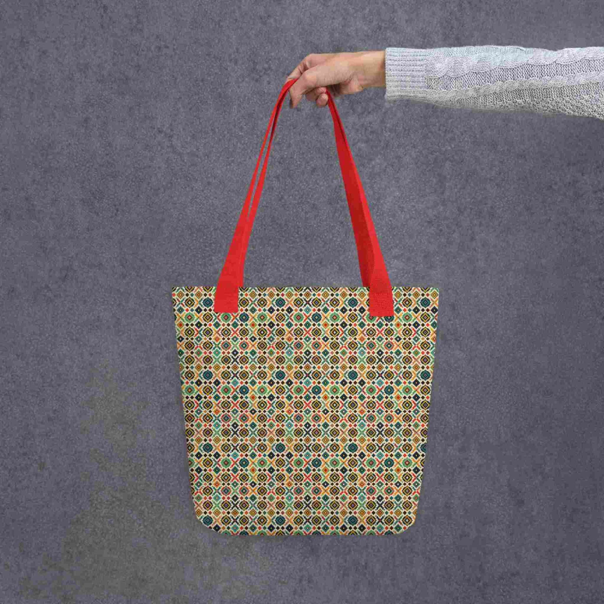 A 15x15 inch tote bag with red handles, which design is an Argentinian inspired diamond pattern, held by a lady's hand. Grey background.