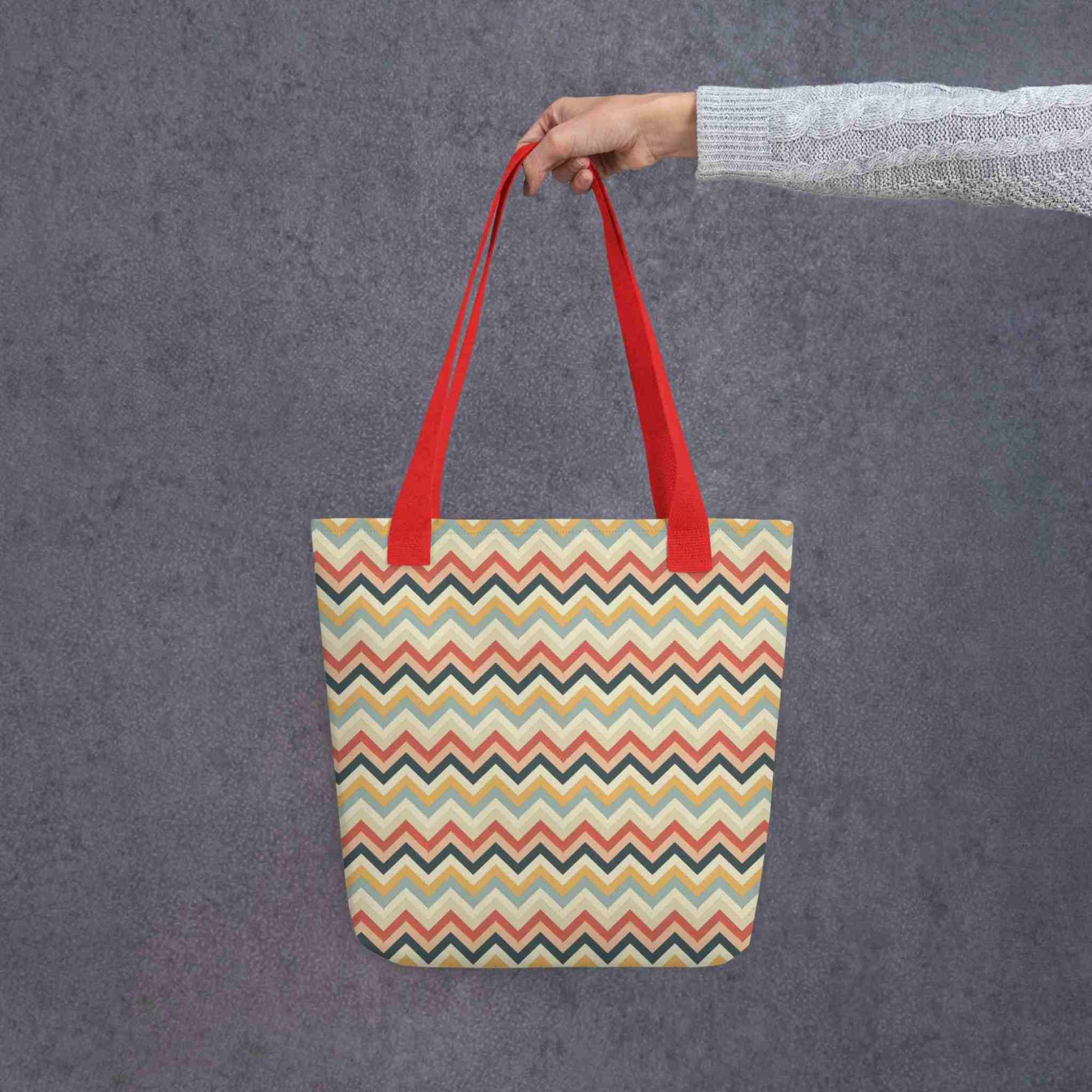 A 15x15 inch tote bag with red handles, in a multicoloured pattern of Zig Zag lines, held in a lady's hand. Grey background.