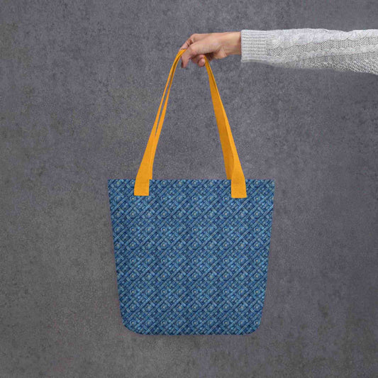Blue tote bag with yellow handles, which features a blue Viking inspired denim pattern, held by a lady's hand. Dimension 15x15 inches. Grey background. 