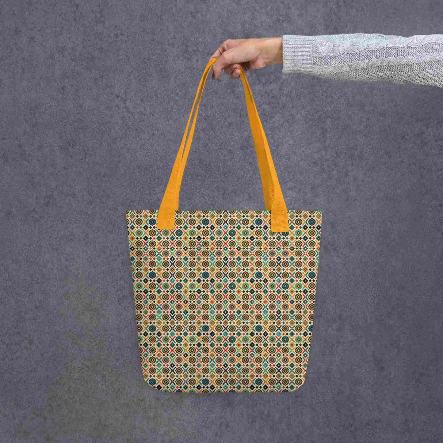 A 15x15 inch tote bag with yellow handles, which design is an Argentinian inspired diamond pattern, held by a lady's hand. Grey background.