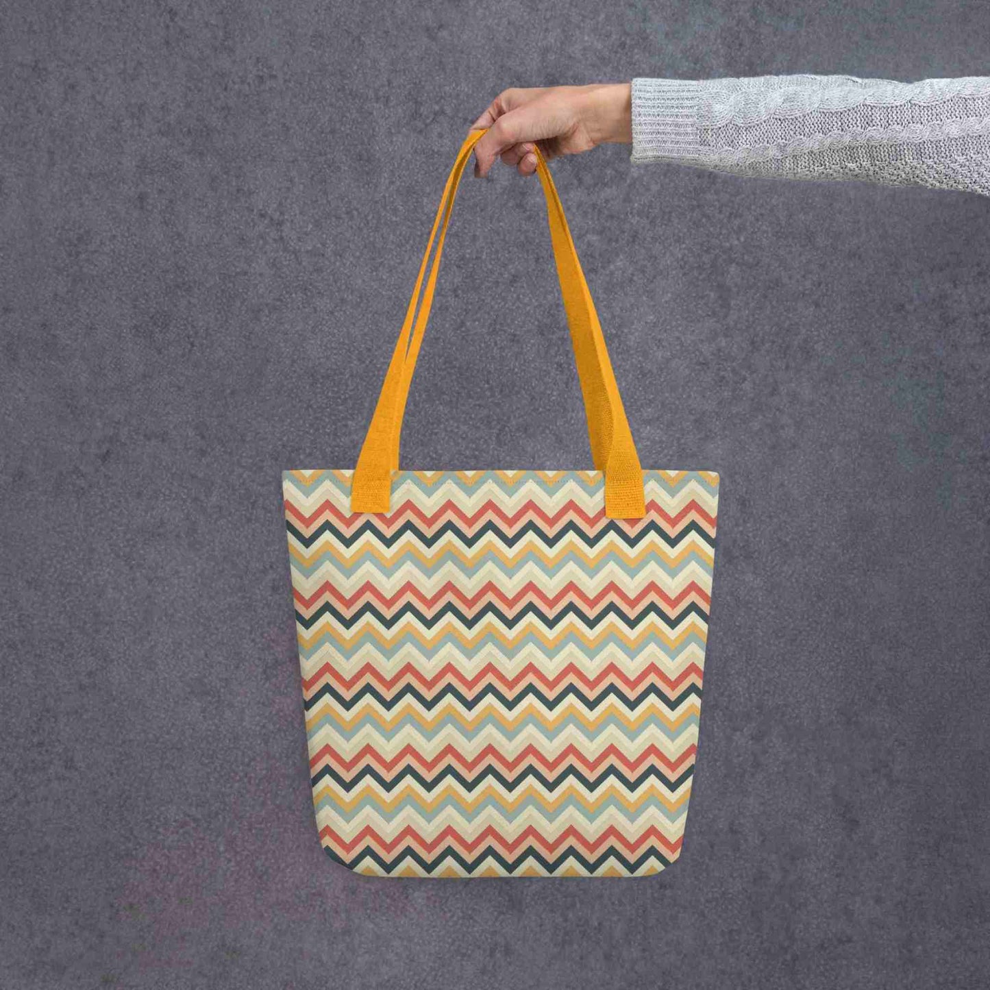 A 15x15 inch tote bag with yellow handles, in a multicoloured pattern of Zig Zag lines, held in a lady's hand. Grey background.