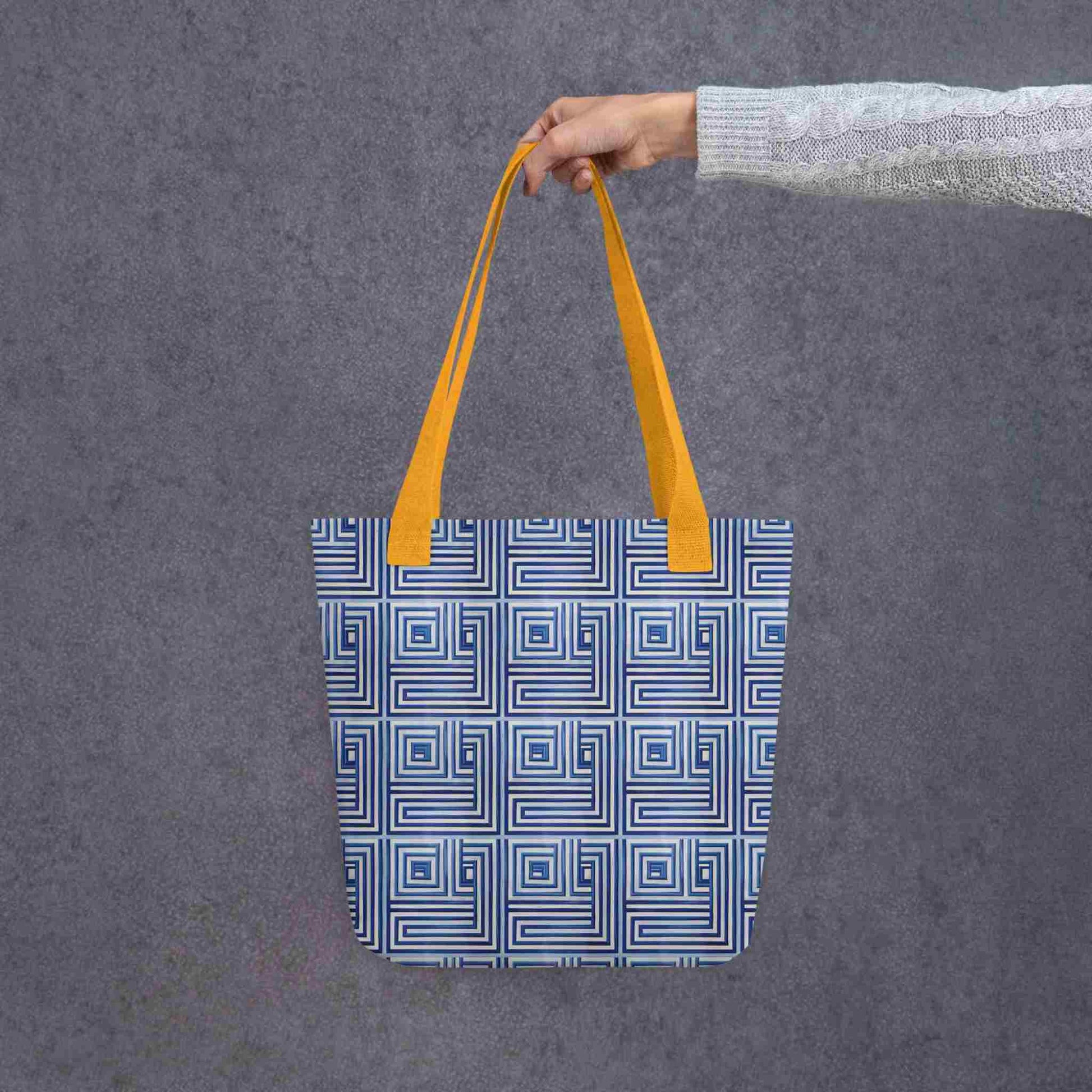 Blue op art tote with yellow handles, which features a blue op art illusion, held in a lady's hand. Dimension is 15x15 inches. Grey background.
