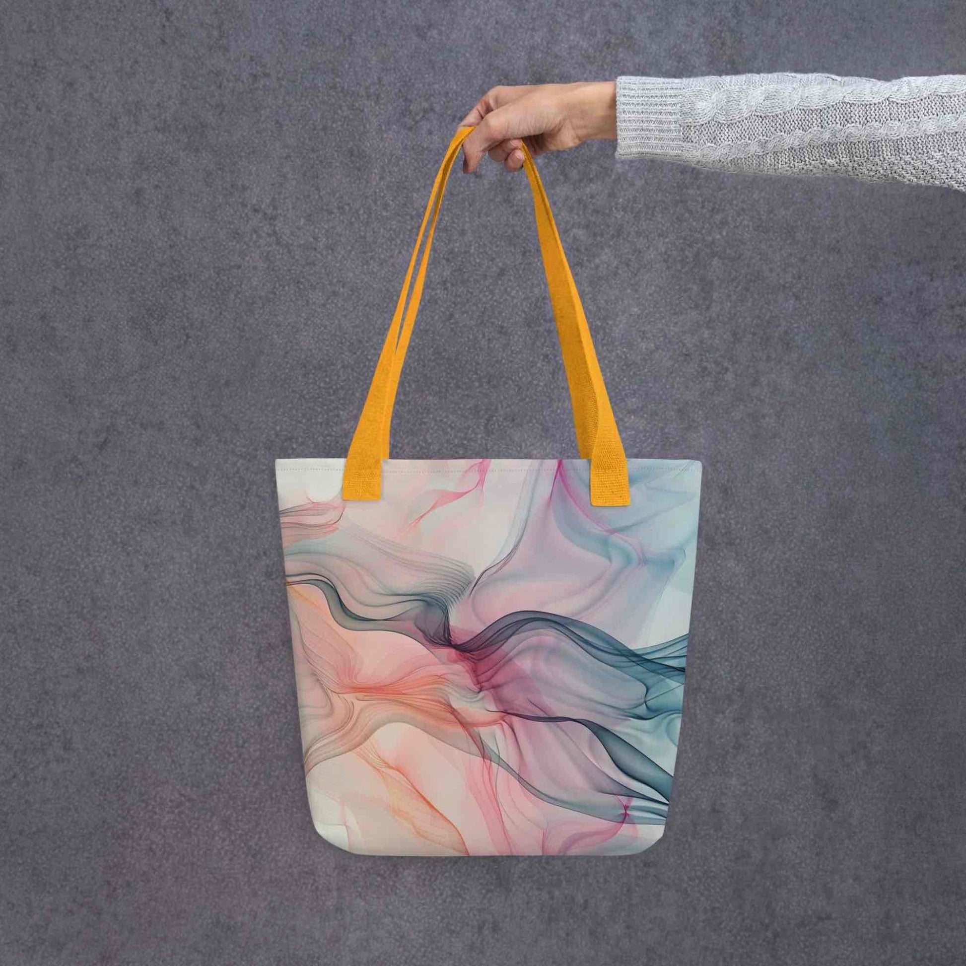 A 15x15 inch tote bag with yellow handles, which design is an abstract art of artistic wavy bluish and pastel lines, held by a lady's hand. Grey background.