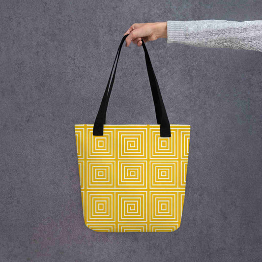 A 15x15 inch tote bag with black handles, with mesmerizing square-patterned op art illusion in a calming yellow, held in a lady's hand. Grey background.